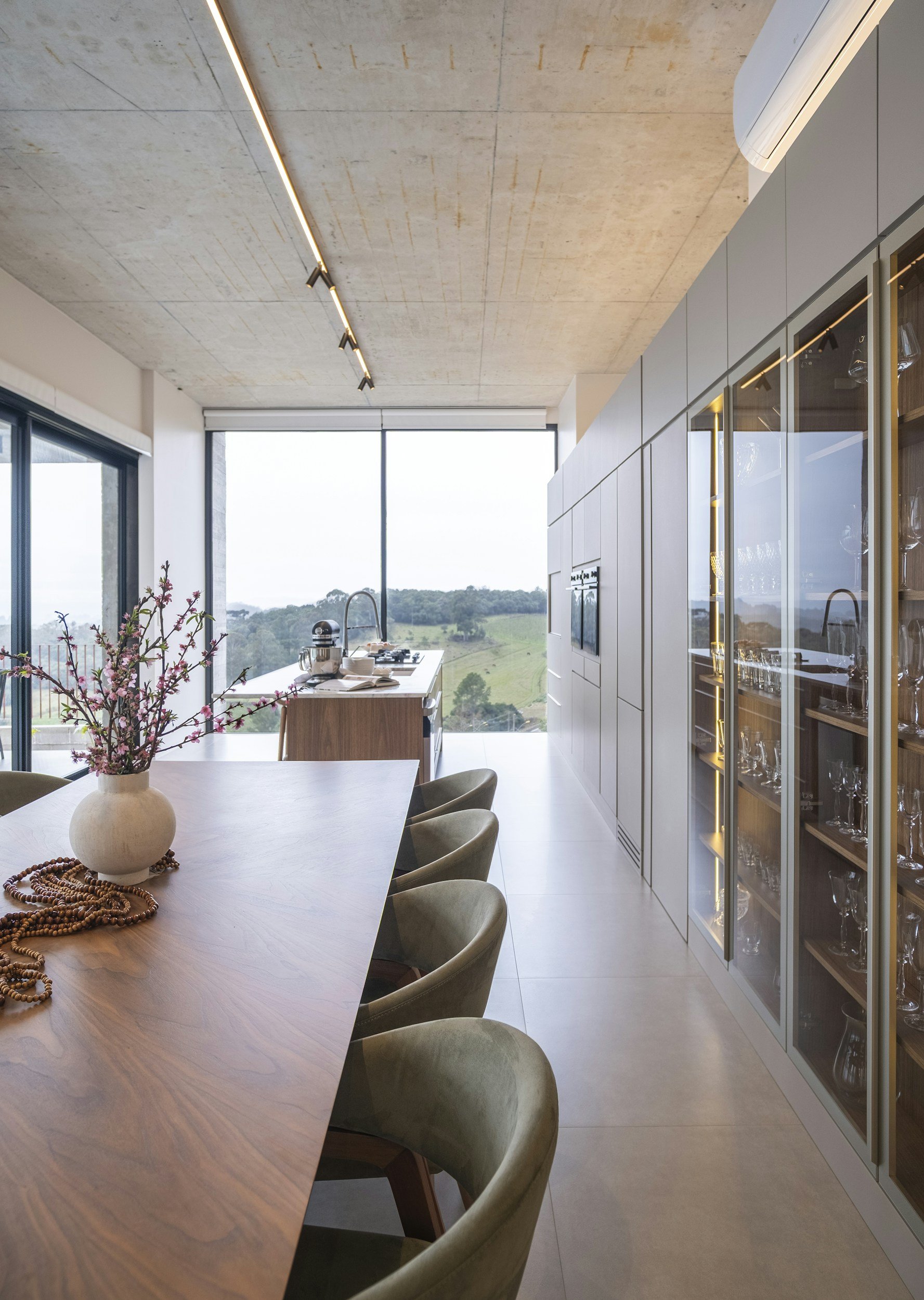 Modern dining area with a wooden table, green upholstered chairs, a vase with pink blossoms, and a large window overlooking a green landscape. Kitchen area with cabinetry and glassware display on the right.