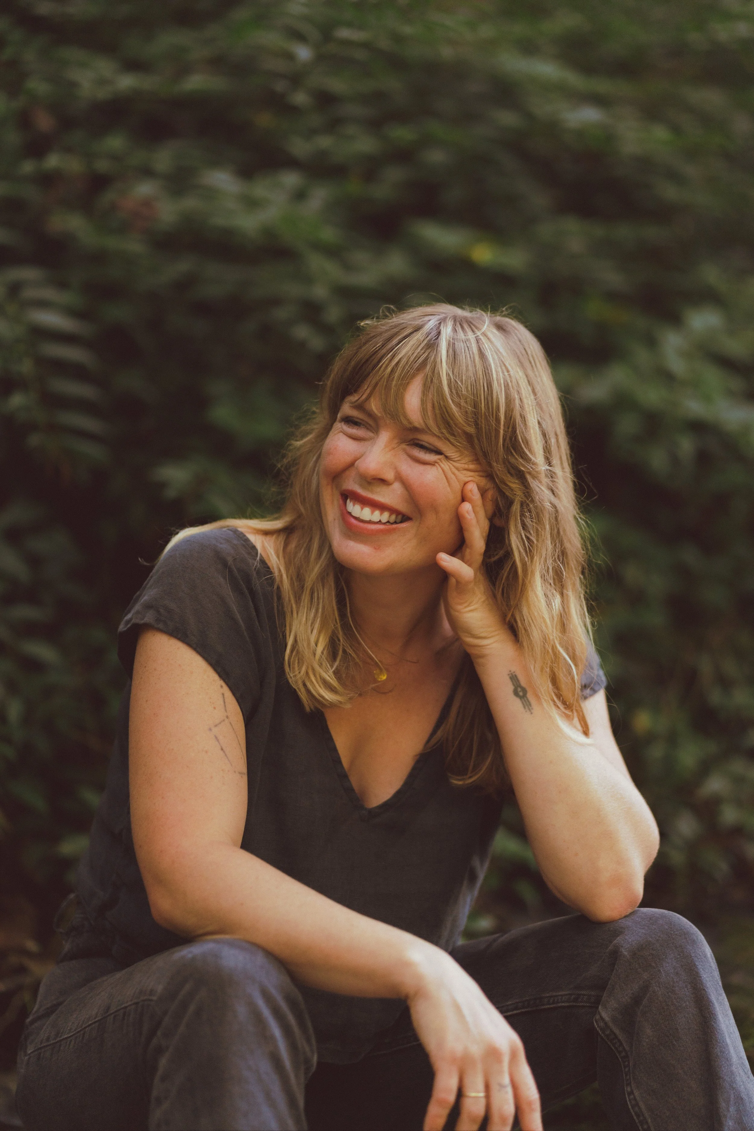 A woman with wavy blonde hair sitting outdoors, smiling and touching her face, with green foliage in the background.