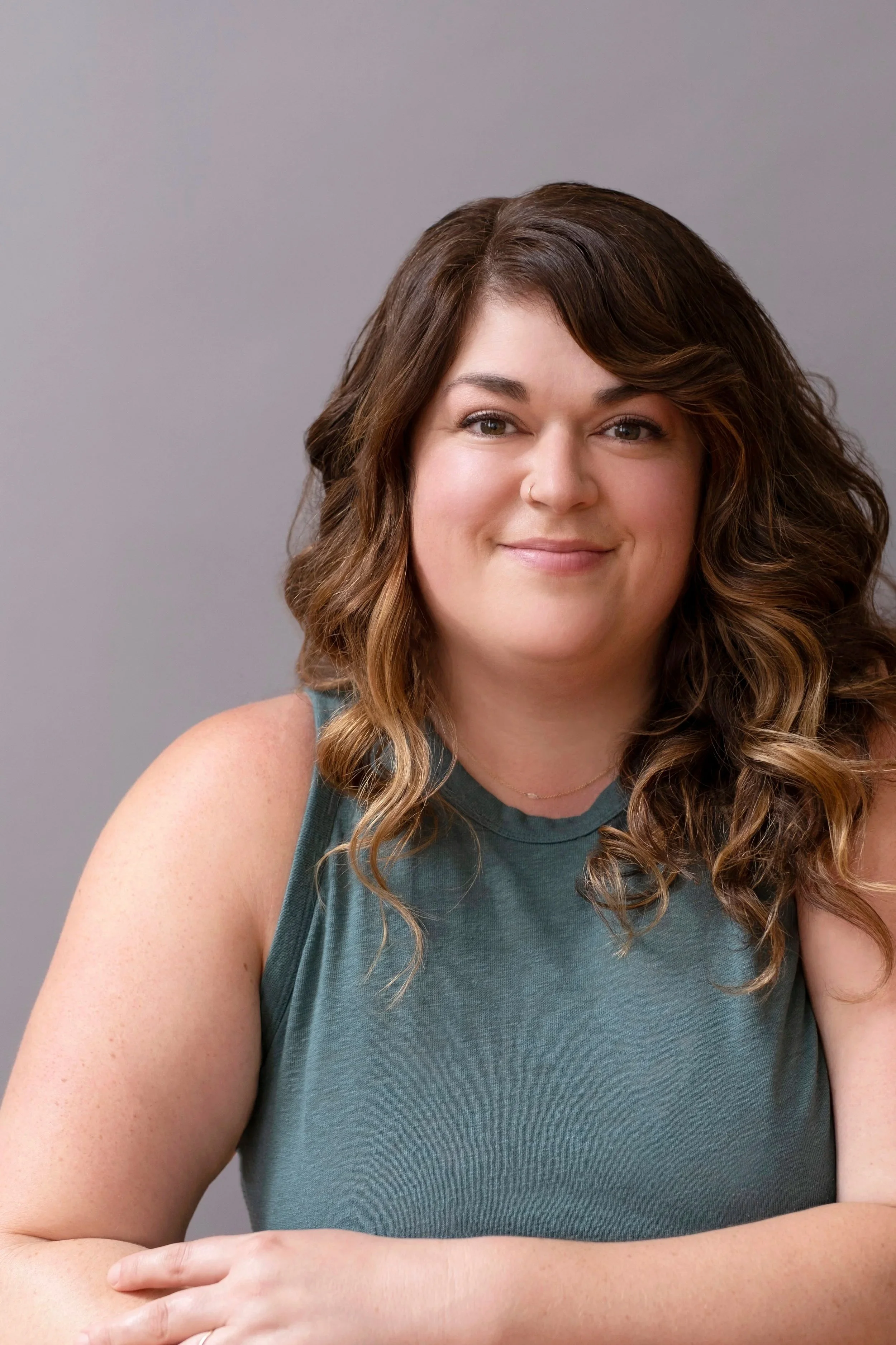 A smiling woman with curly brown hair, wearing a sleeveless teal top, resting her arm on a surface against a plain gray background.