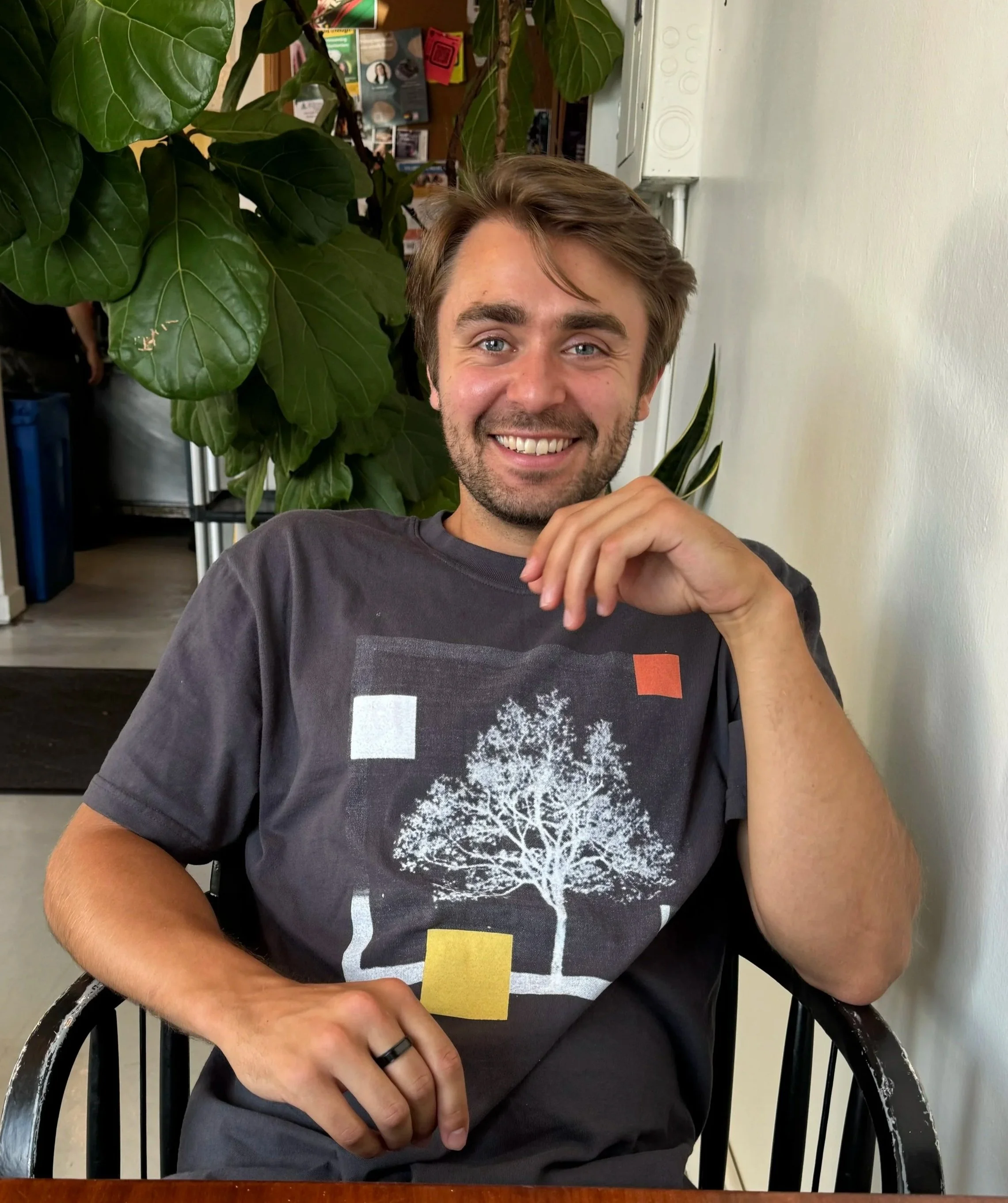 A young man with brown hair and beard smiling while sitting at a table, wearing a black t-shirt with a white tree graphic and colorful squares, with large green leaves behind him.