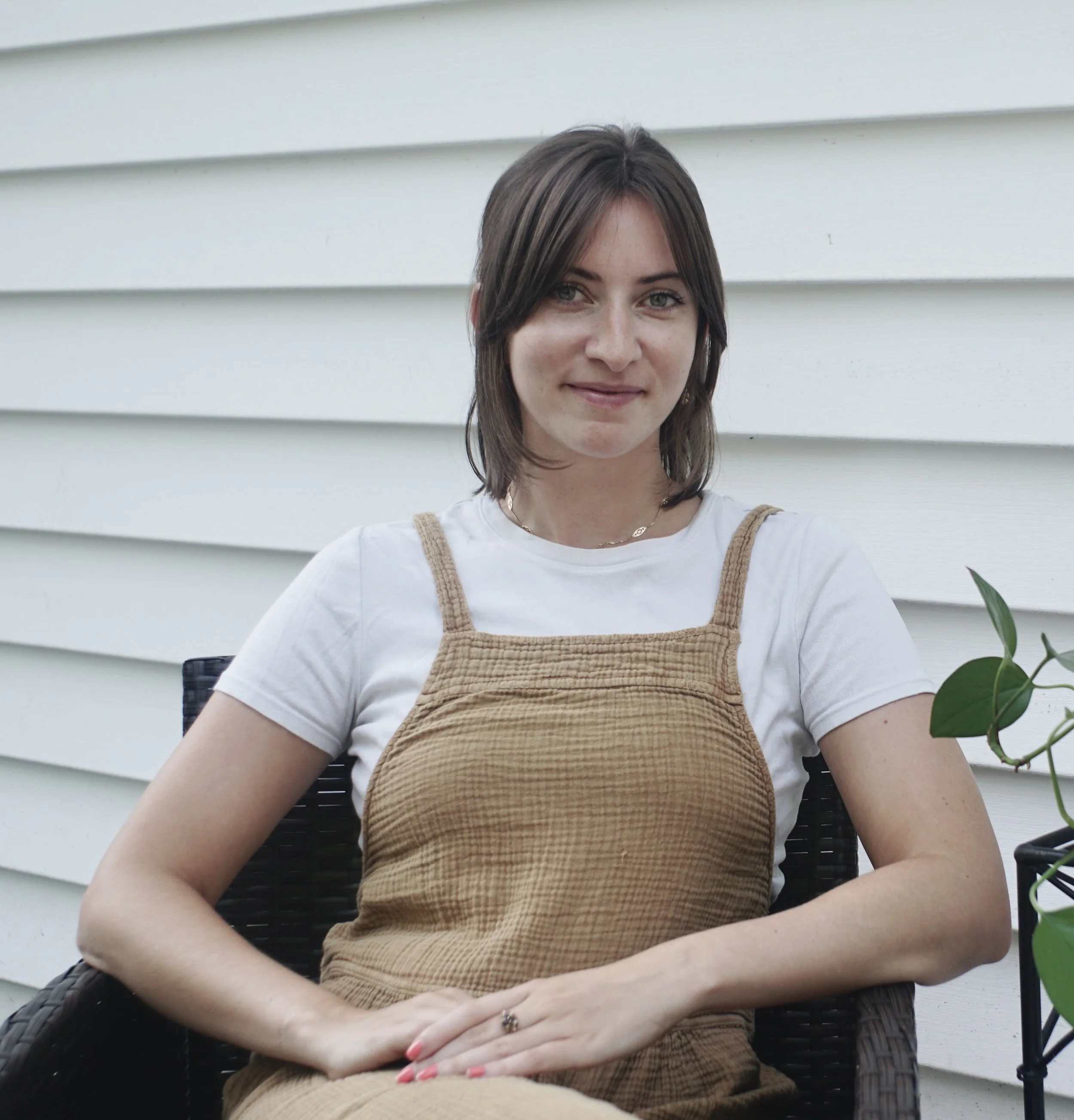 Young woman with shoulder-length brown hair smiling, sitting outdoors in front of white horizontal siding, wearing a white T-shirt and brown overalls, with a small potted plant nearby.
