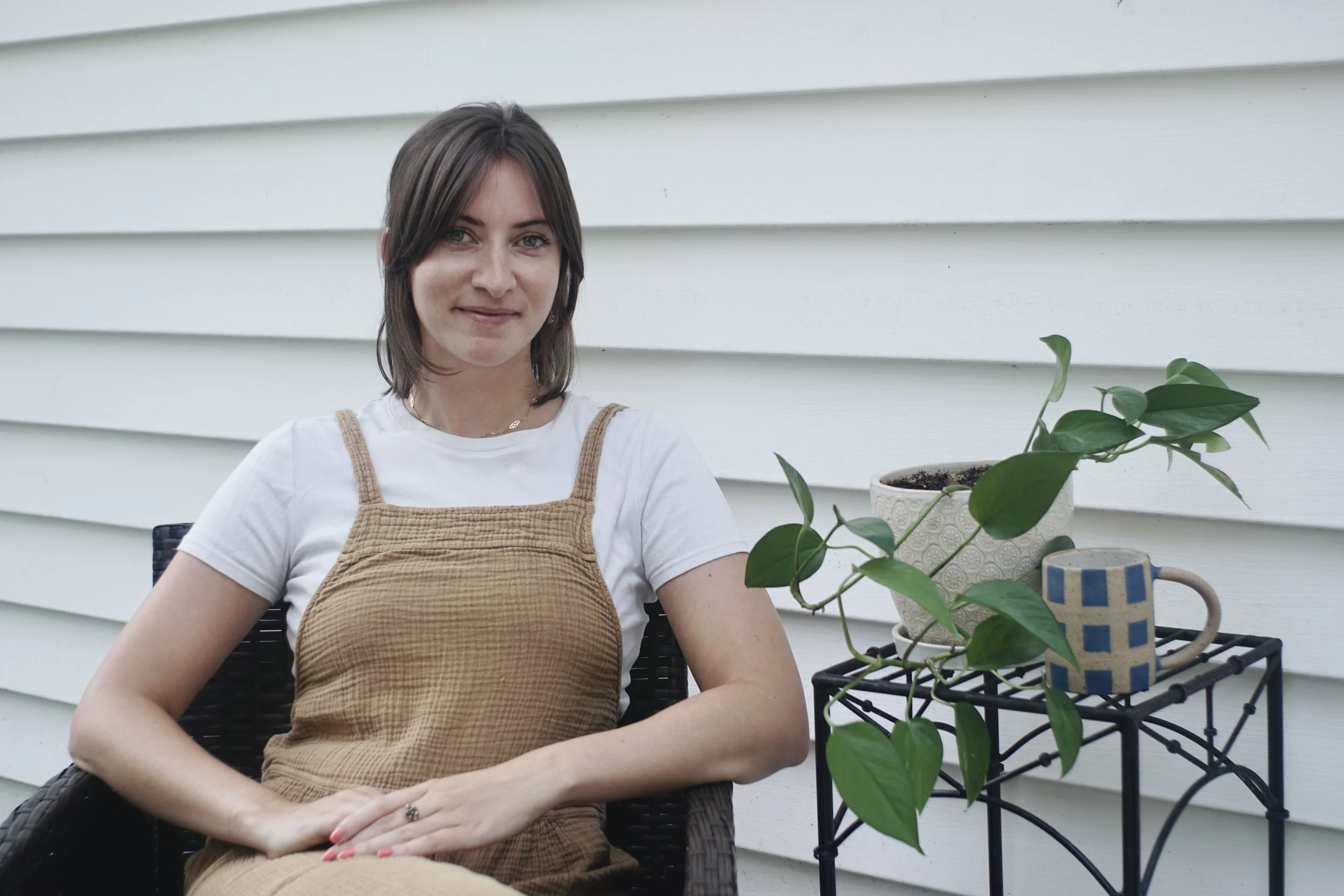 Young woman with brown hair, wearing a white t-shirt and tan dress, sitting on a black woven chair outside next to a black metal plant stand with a potted plant and a blue and beige mug, against a white wooden house siding background.