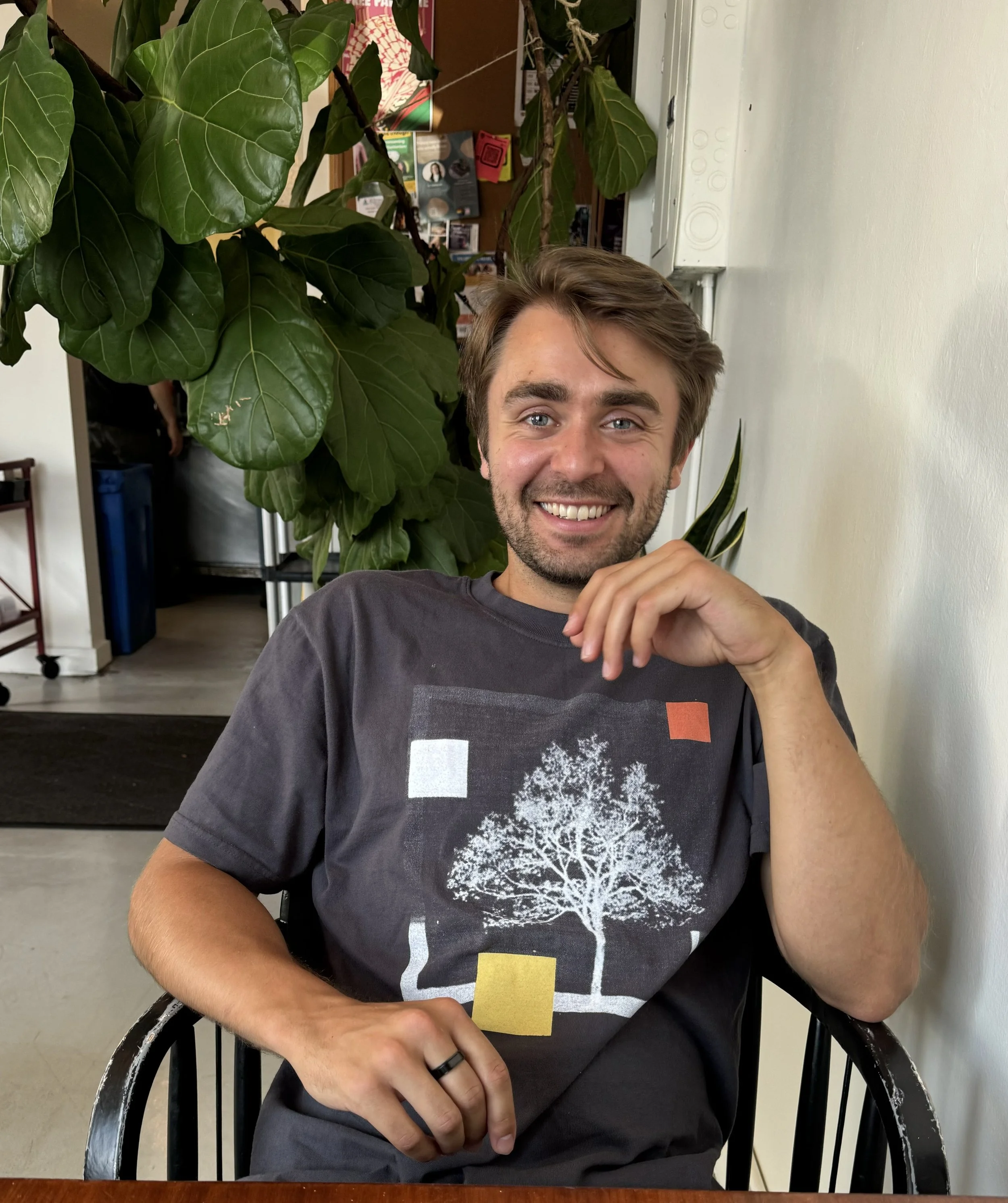 A young man with light brown hair, a beard, and blue eyes sitting at a table, smiling. He is wearing a dark gray T-shirt with an abstract tree and colorful blocks on the front. There is large green leafy plant behind him and a white wall to his right.