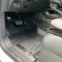 Car interior with black rubber floor mat, dashboard, and driver side foot pedals.