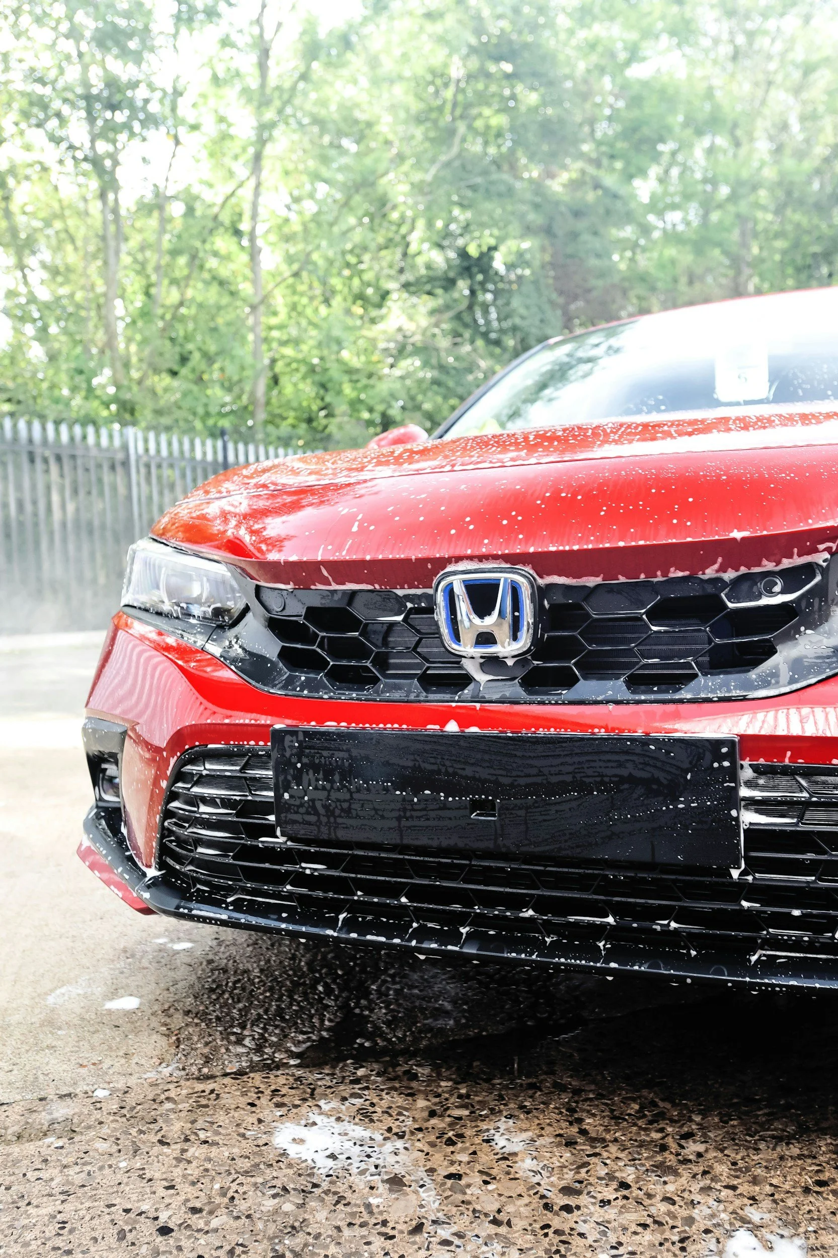 Front view of a red Honda car with soap suds on the surface after washing, outdoors on a driveway, with trees and a fence in the background.