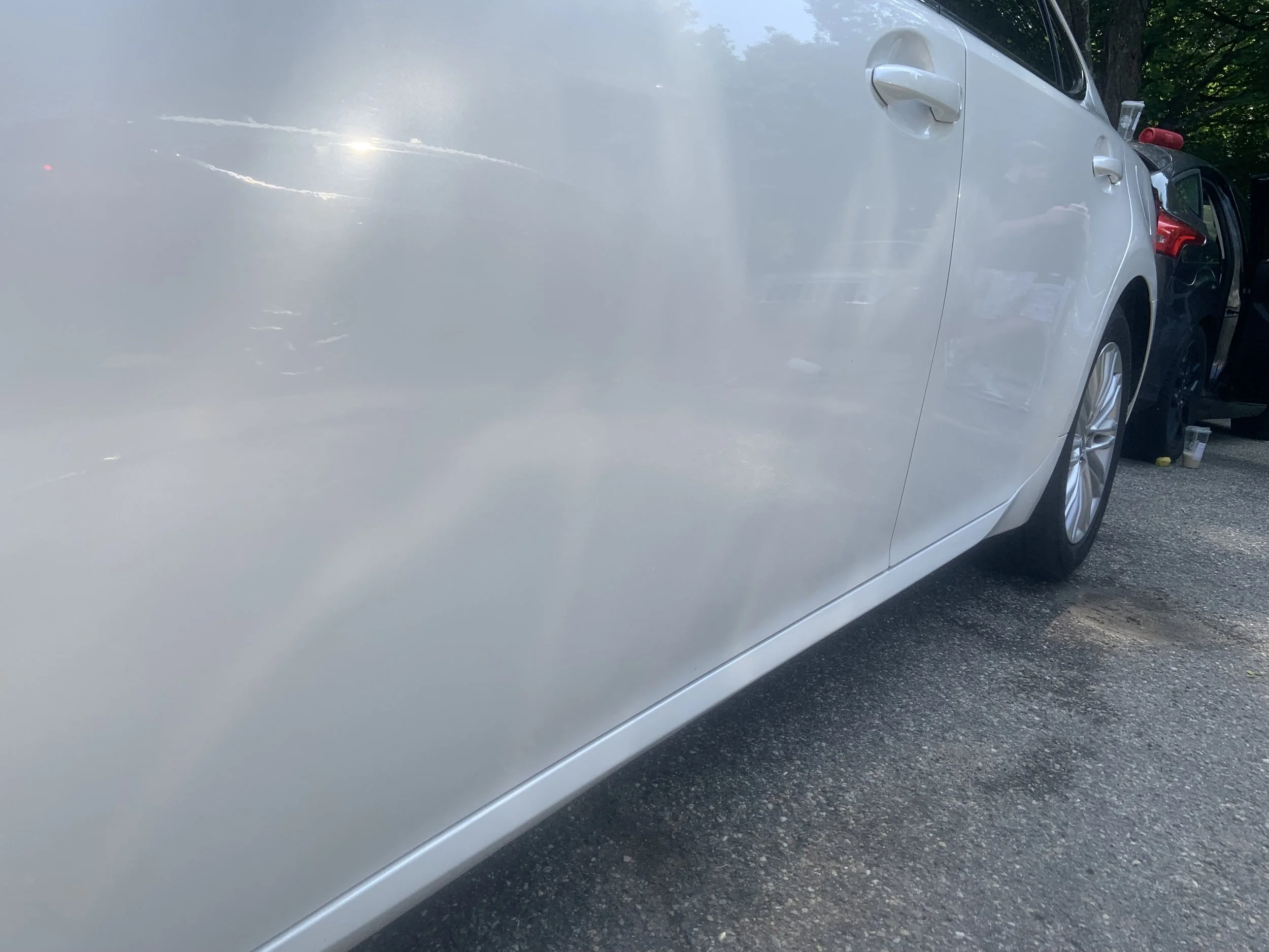 Close-up of a white car with visible scratches and dents on the side panel, parked on a paved surface.