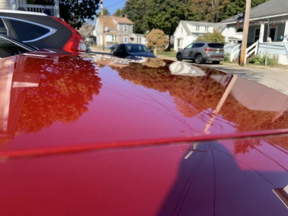 Close-up of a shiny red car roof reflecting trees, power lines, and houses in a neighborhood street.