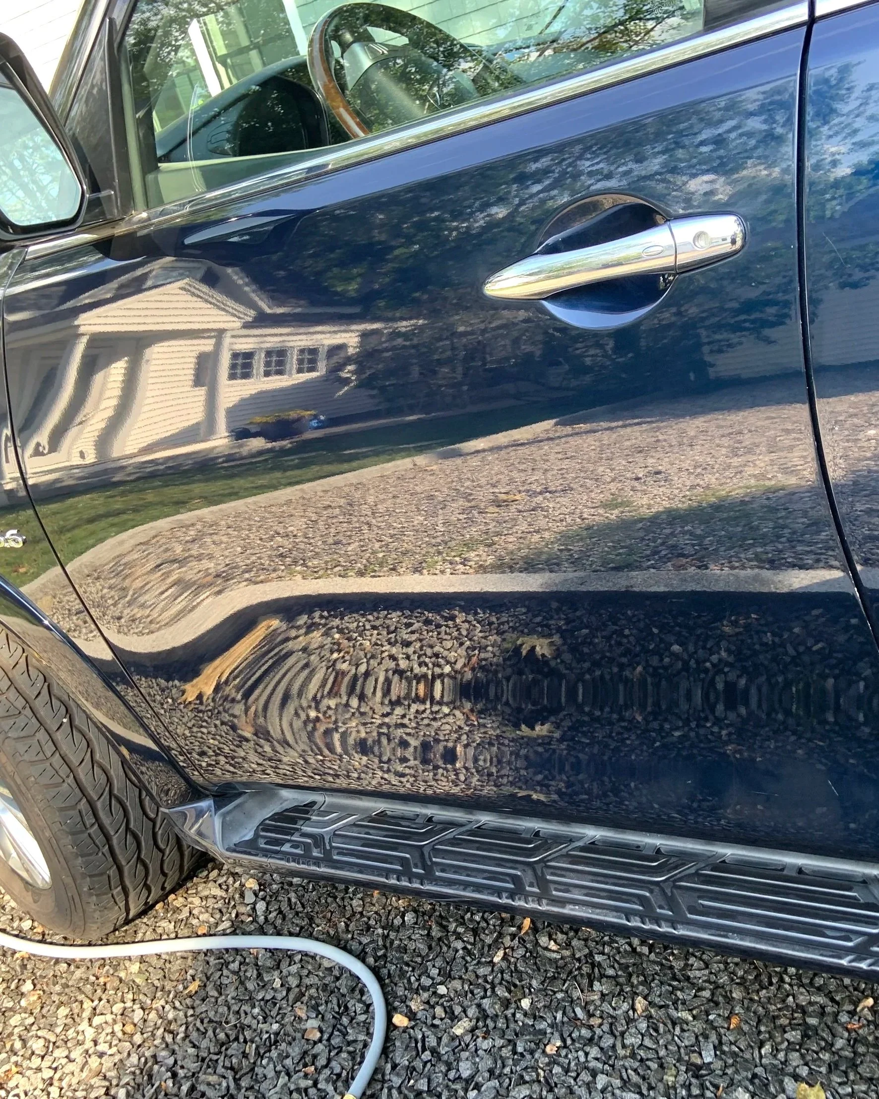 Close-up of a black car's side door with a chrome handle, reflecting a house and trees, parked on a gravel driveway.