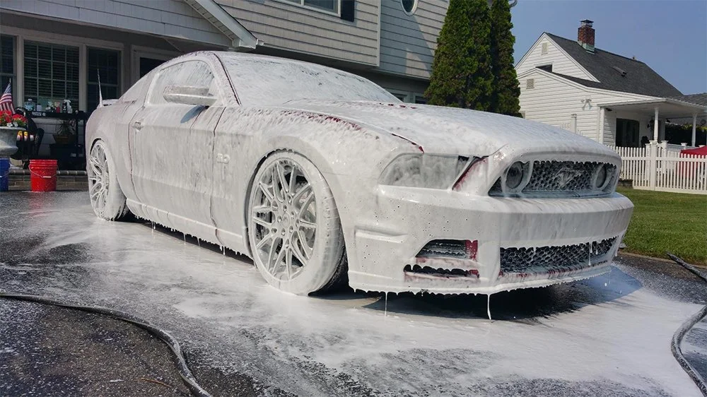 A white sports car covered in white foam during a car wash outside a house with a yard and neighboring houses.