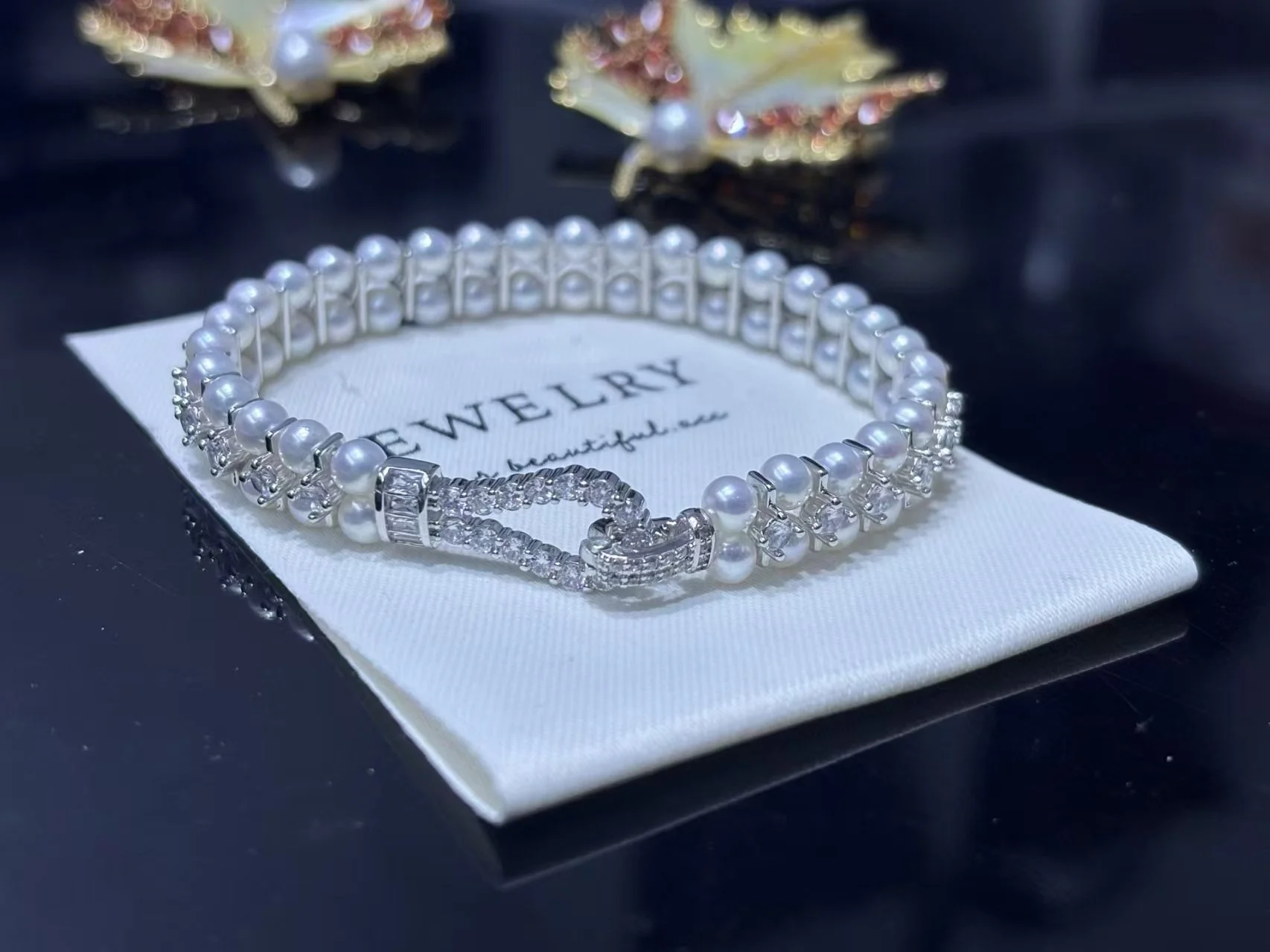 A pearl bracelet with a clasp, placed on a white jewelry cloth with the words "Jewelry" and "beautiful" written on it, alongside decorative earrings in the background.