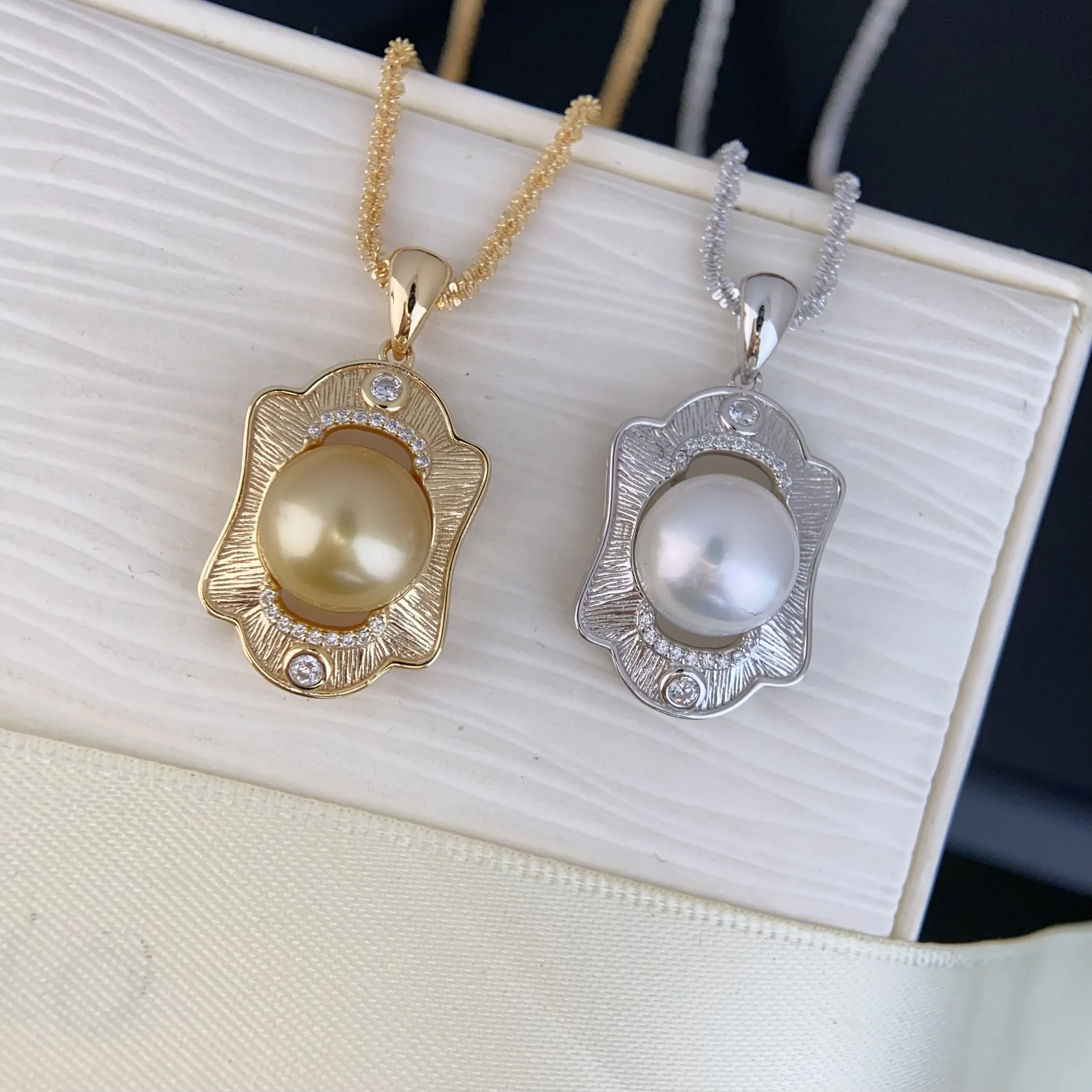 Two pendant necklaces, one gold and one silver, each with a large pearl in the center surrounded by small diamonds, displayed on a white jewelry stand.