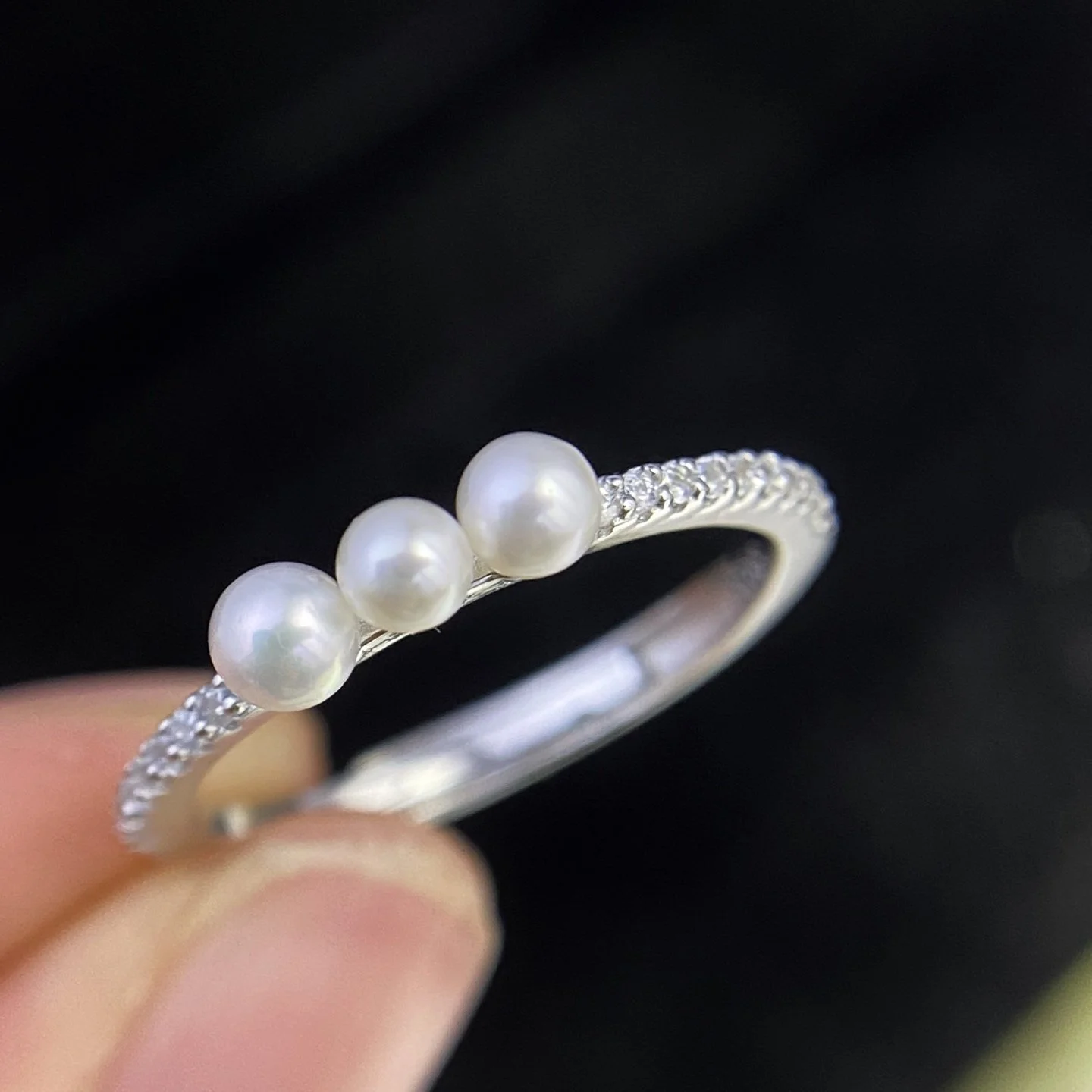 Silver ring with four white pearls and small diamonds on the band, held by a person's fingers against a black background.