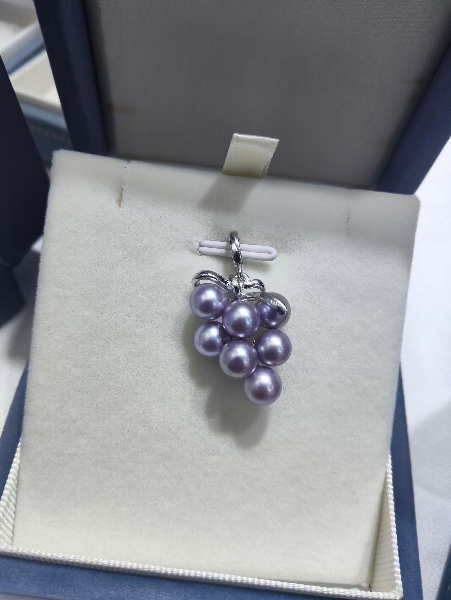 A pendant in a jewelry box shaped like a bunch of purple grapes with pearl-like beads glued together, hanging on a metallic bail.