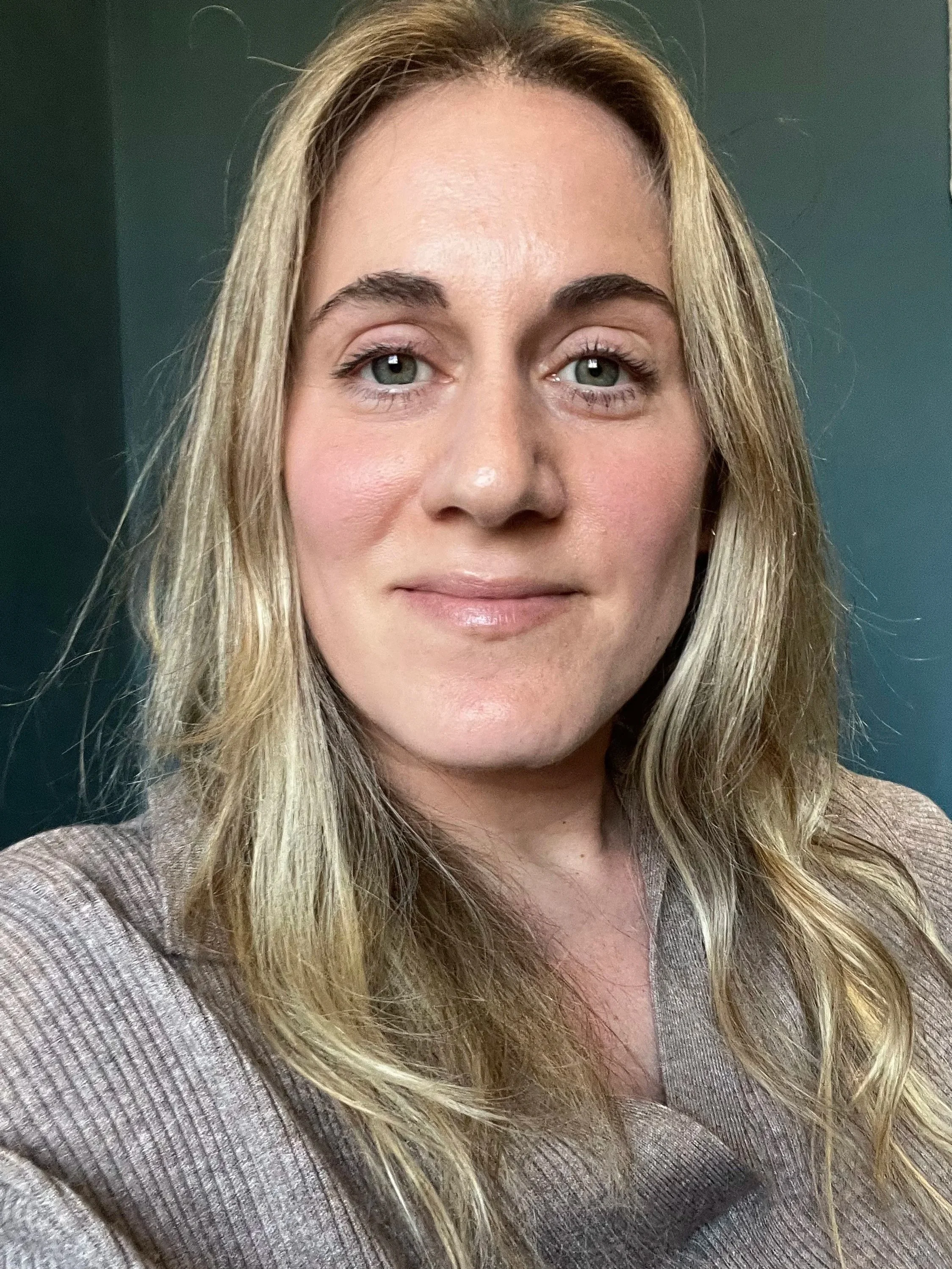 Close-up of a female counsellor with long blonde hair, light skin, and wearing a brown top, standing against a greenish background which is her therapy room.