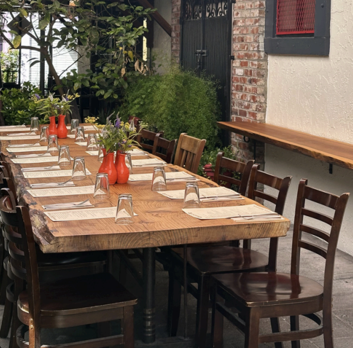 A long wooden dining table set with glasses, knives, and cream-colored napkins, with red vases containing flowers as centerpieces, in an outdoor patio area with chairs, greenery, and brick and stucco walls.