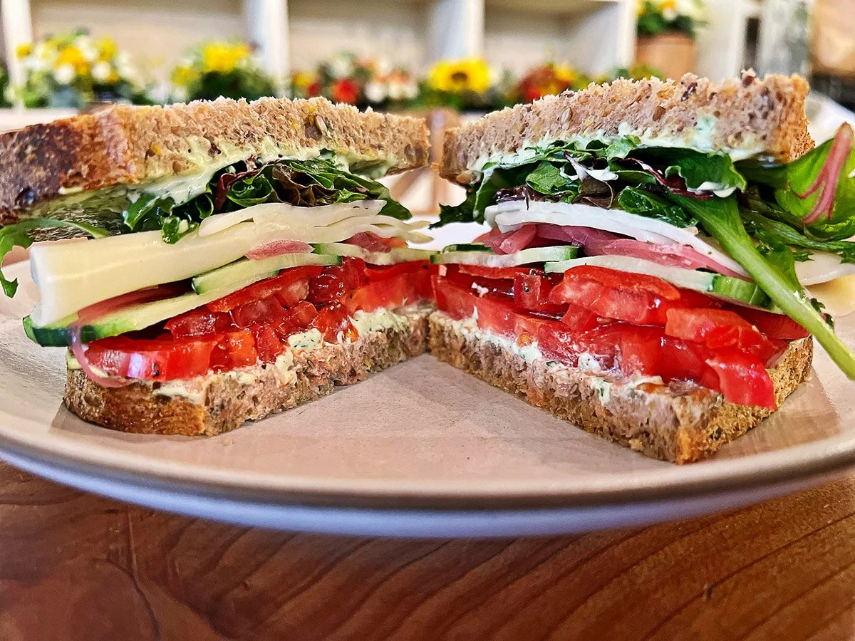 Cross-section of a veggie sandwich with tomatoes, cucumbers, cheese, leafy greens, and whole grain bread on a plate.