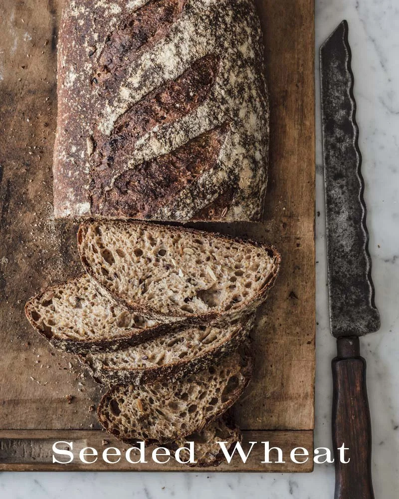 A loaf of seeded wheat bread partially sliced on a wooden cutting board with a bread knife beside it.