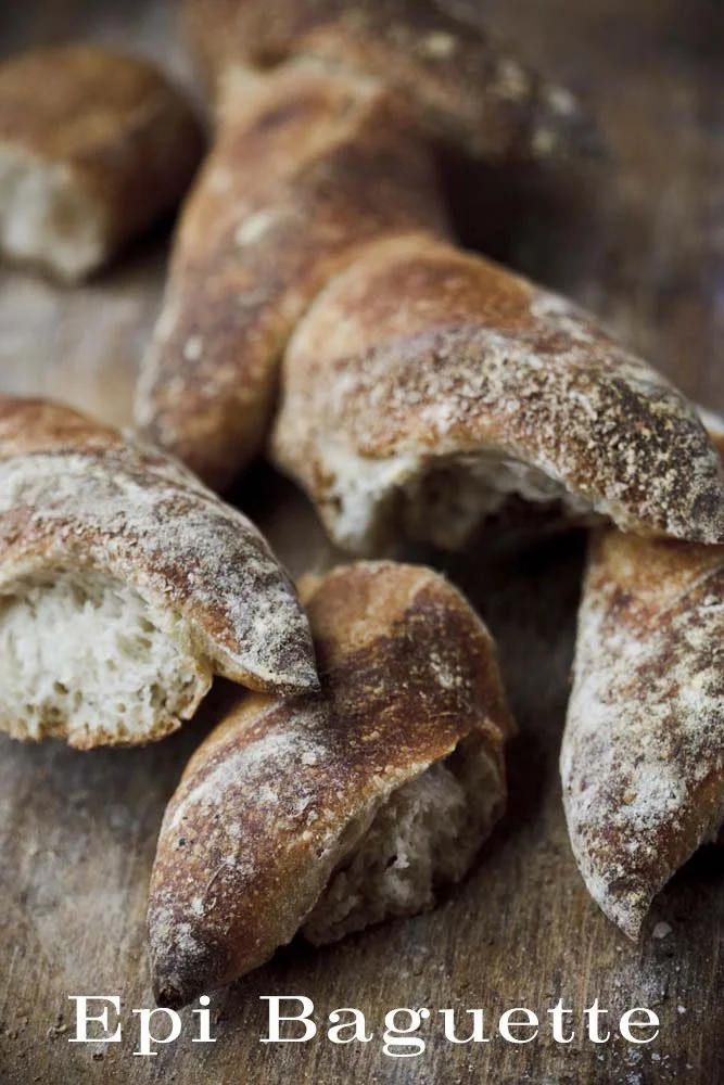 Close-up of baked croissants with flaky, crispy exterior, some broken open showing soft interior, on a wooden surface, labeled "Epi Baguette".