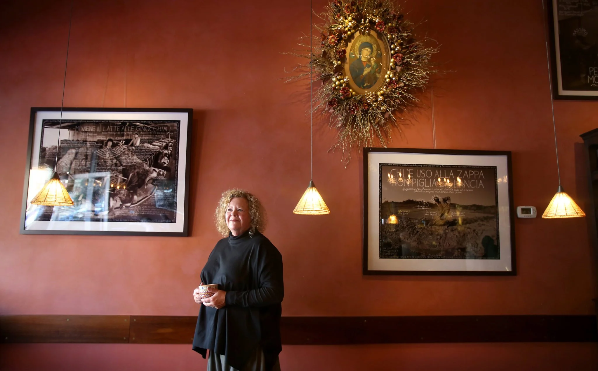A woman with curly blonde hair wearing a black top holds a cup while standing inside a restaurant or café with rust-colored walls, decorated with framed black and white photographs, hanging pendant lights, and a Christmas wreath with gold and red ornaments surrounding a religious icon.