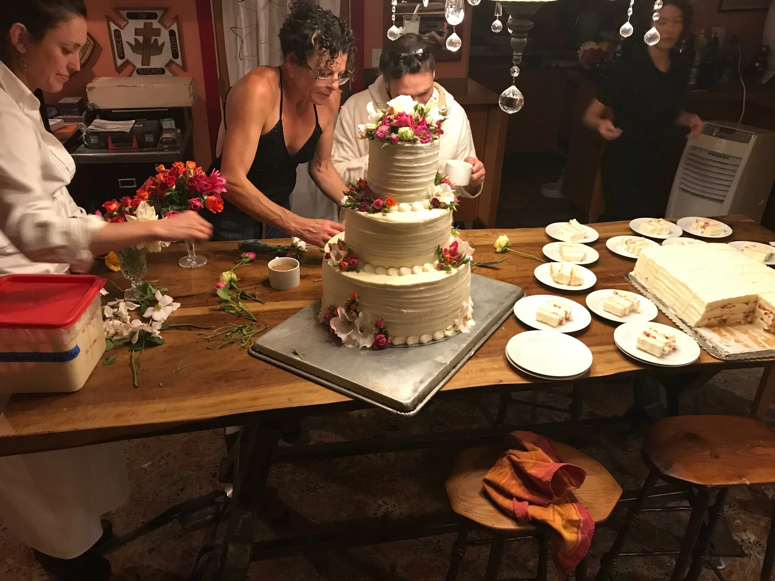 People gather around a table with a large three-tiered wedding cake decorated with flowers, surrounded by plates of cake slices and floral arrangements, in a cozy indoor setting.