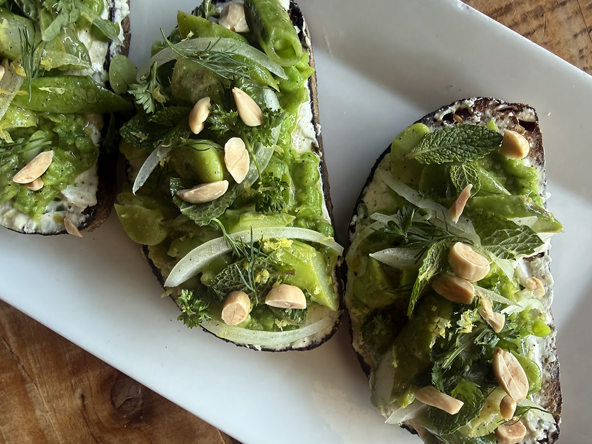 Two slices of avocado toast topped with fresh herbs, chopped green vegetables, and slivered almonds, served on a white plate.