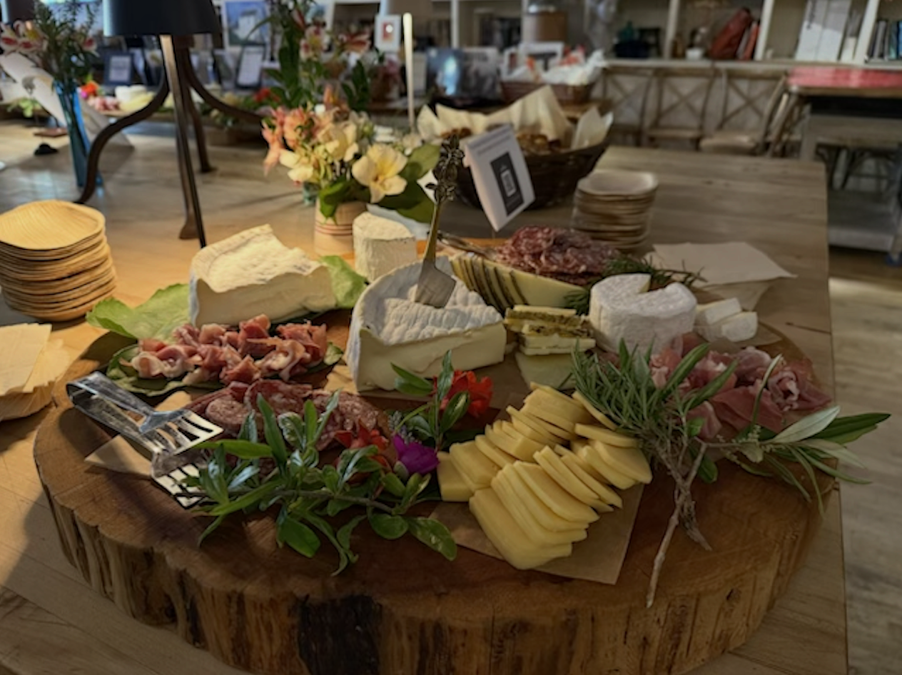 Cheese and charcuterie platter with assorted cheeses, cured meats, fresh herbs, and flowers on a wooden serving board in a rustic restaurant setting.