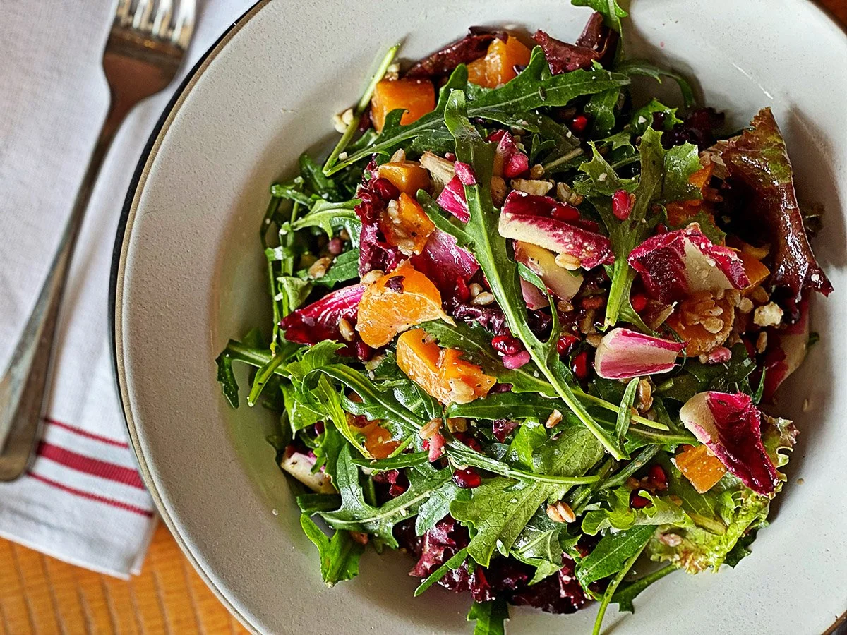 A bowl of mixed salad with arugula, radicchio, orange segments, chopped nuts, and seeds.
