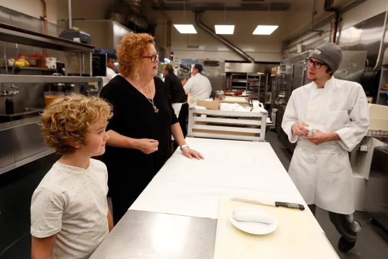 A woman and a young boy in a kitchen talking to a chef wearing a white coat and a gray beanie.