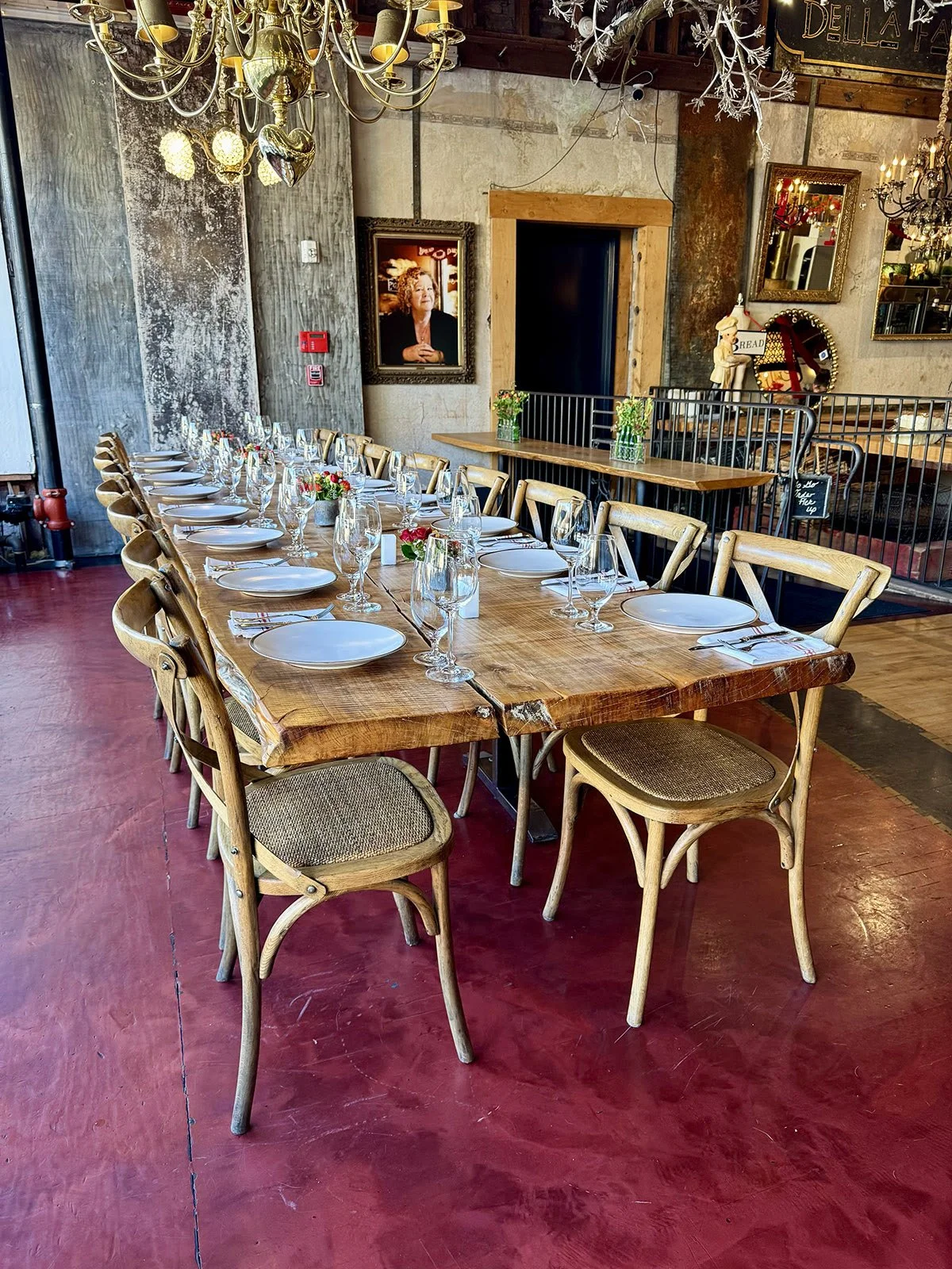 A long wooden dining table is set for a meal with white plates, wine glasses, and small floral centerpieces. The room has rustic decor with chandeliers and framed pictures on the walls.