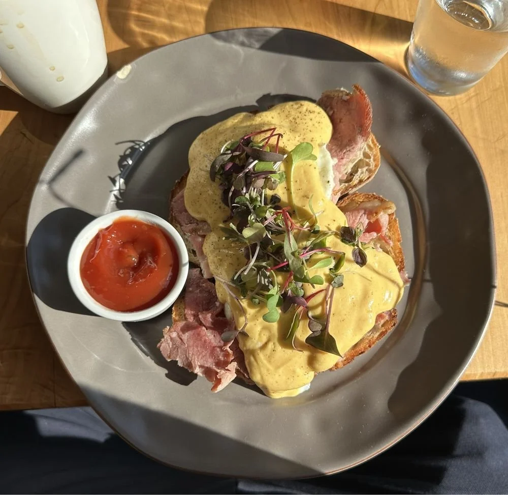 Plate of eggs Benedict with microgreens, served with a side of tomato sauce, water, and a bottle of creamy beverage on a wooden table.