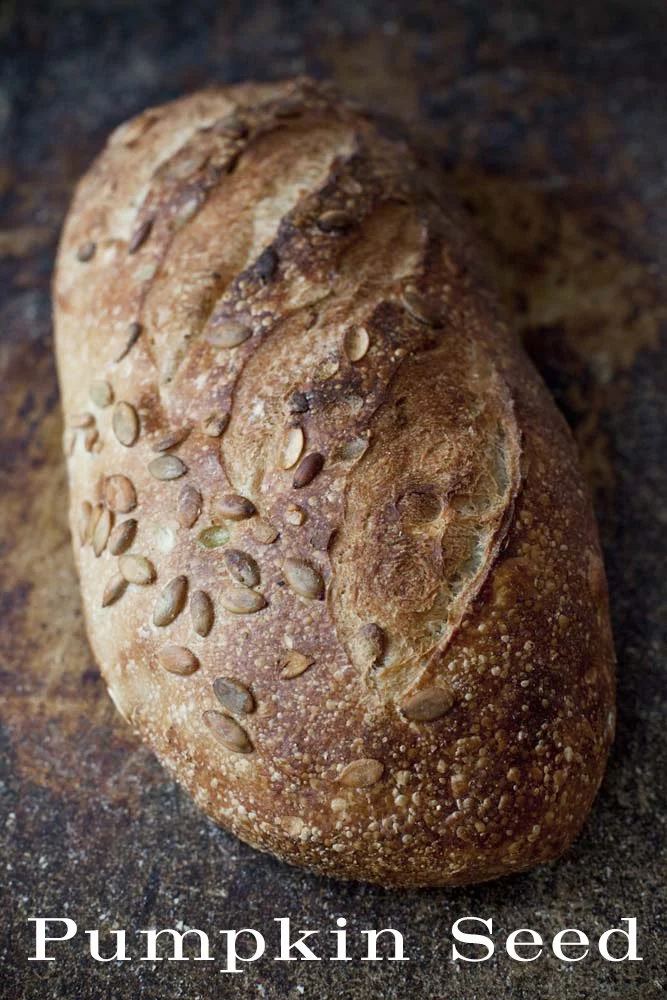 A loaf of bread topped with pumpkin seeds on a dark surface.