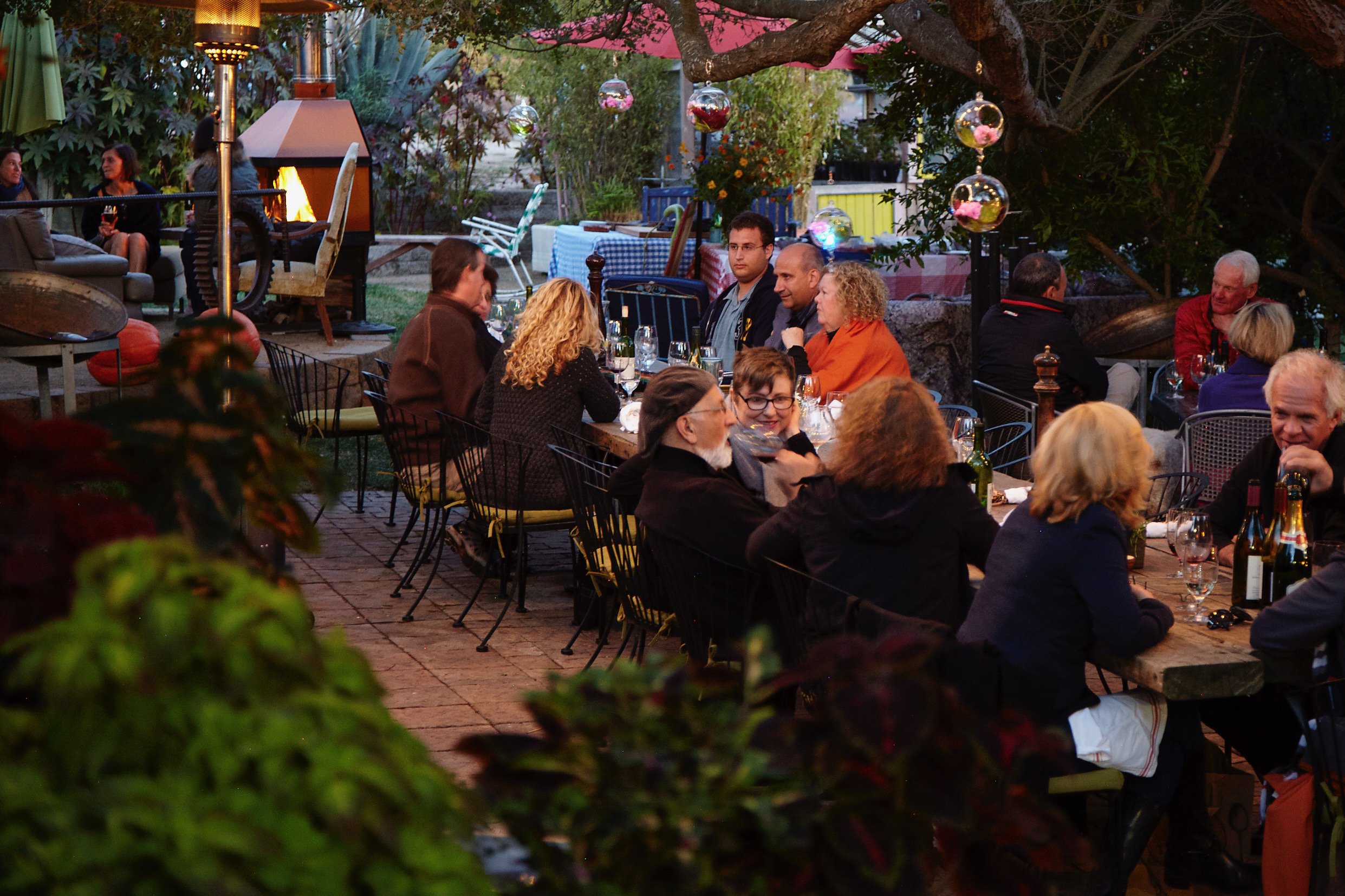 People dining outdoors at a long table in a garden-like setting with hanging glass orbs and a fire pit in the background.
