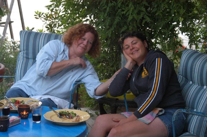 Two women sitting outdoors at a table, one with curly red hair wearing a light blue shirt and the other with dark hair wearing a dark hoodie, smiling and touching hands, with blue cushions and greenery in the background.