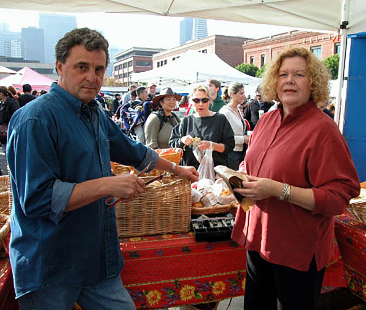 Two people standing at an outdoor market, one man holding a basket and the woman holding a piece of paper, with a crowd and market stalls in the background.