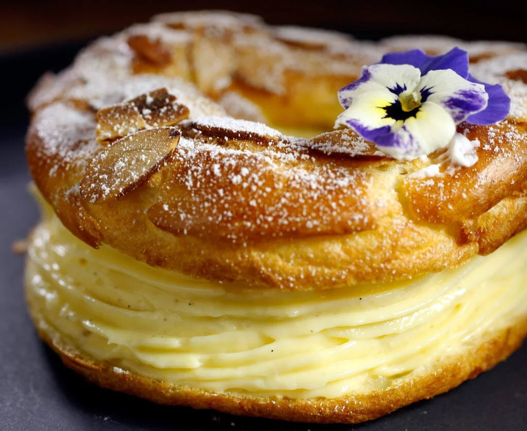 Close-up of a layered pastry dessert topped with powdered sugar, sliced almonds, and a purple and white edible flower.