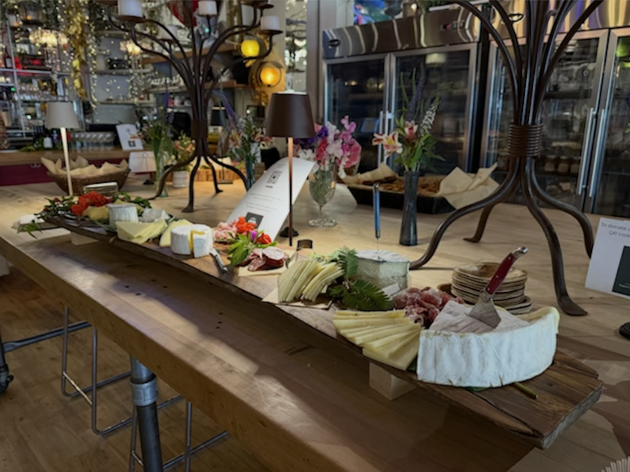A wooden serving board displaying various cheeses, sliced meats, and garnishes on a table in a cozy decor setting with flowers and lamps.
