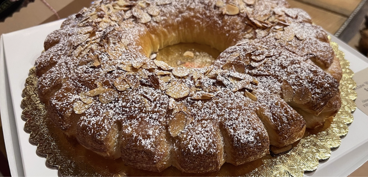 A ring-shaped pastry topped with sliced almonds and powdered sugar, displayed on a gold-colored decorative tray.