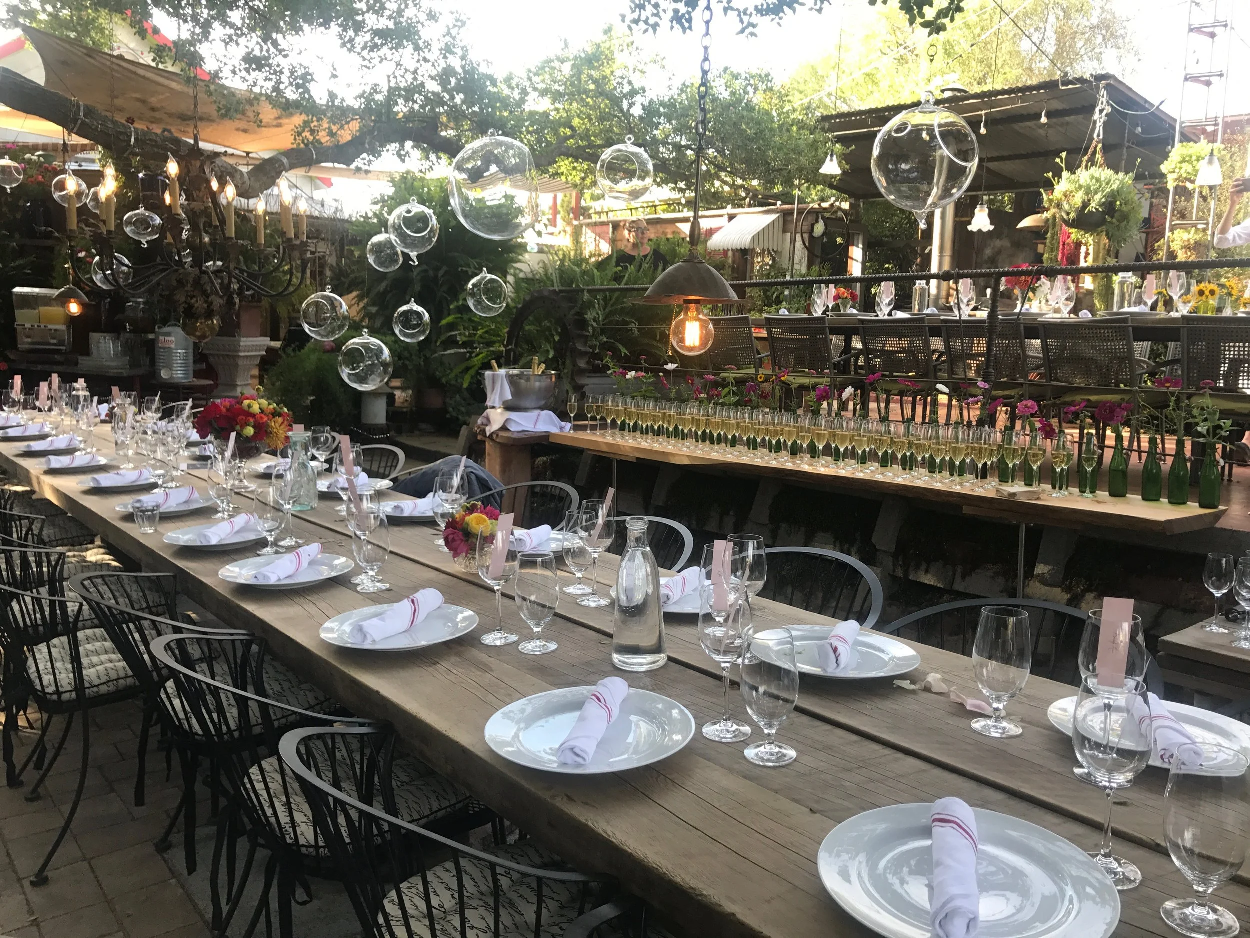 Decorated outdoor dining area with a long wooden table set with white plates, glasses, and napkins, surrounded by black chairs, and adorned with flowers and hanging glass ornaments.