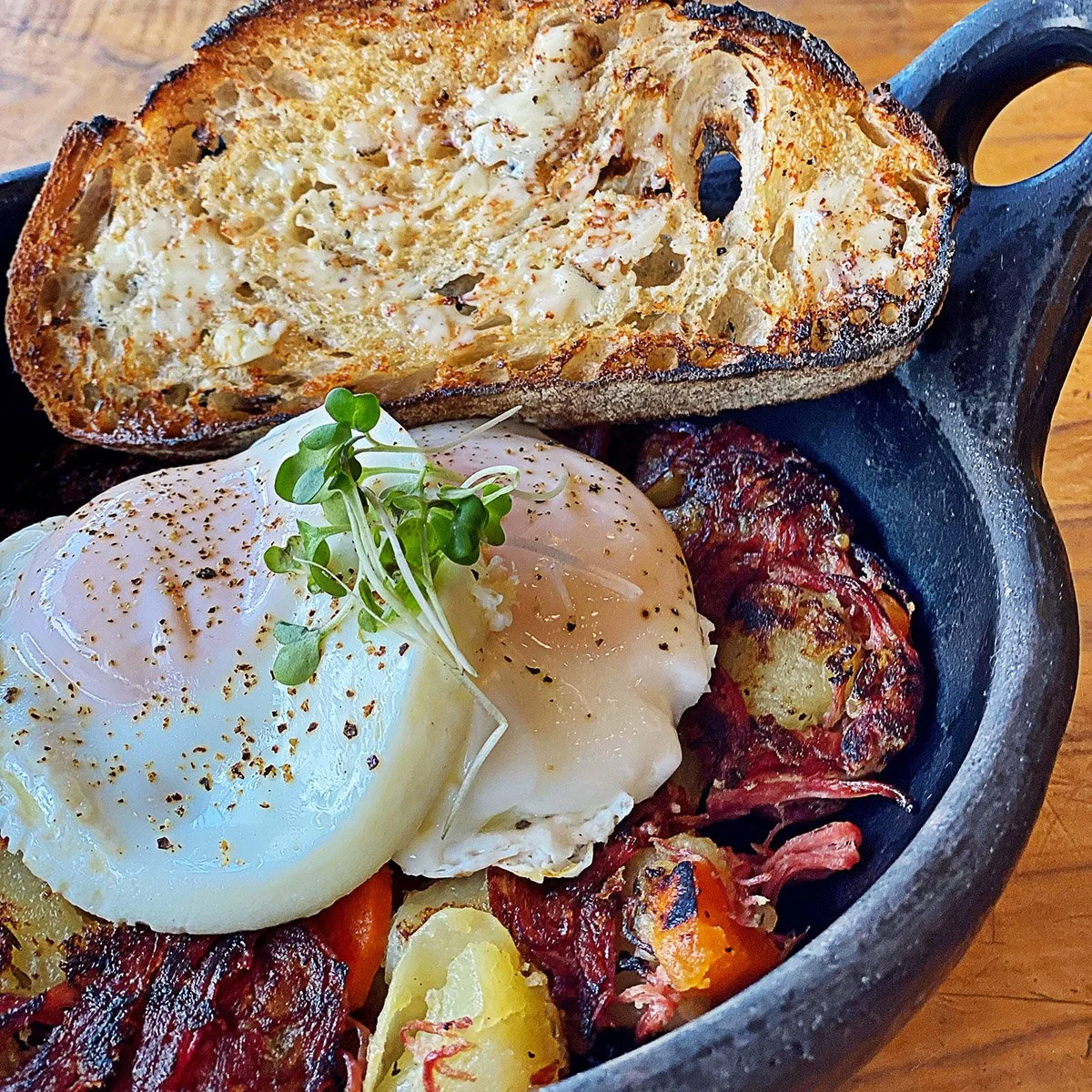 Cast iron skillet with toasted bread, poached egg garnished with microgreens, roasted meat, and vegetables.