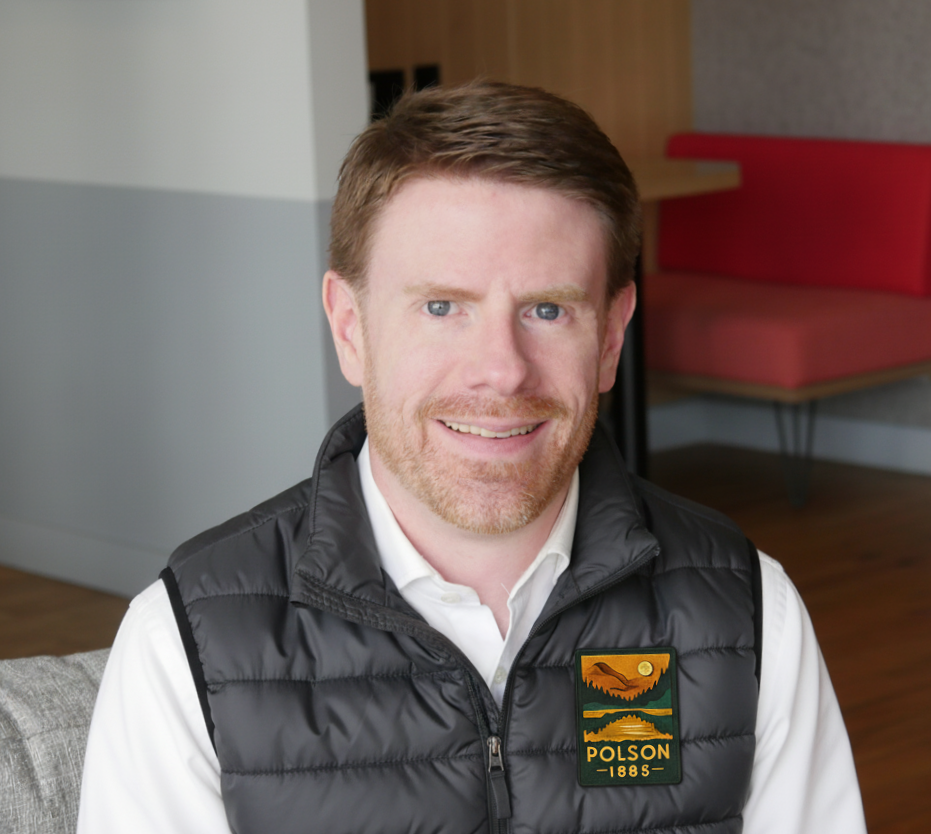 A man with light skin, red hair, and a beard sitting on a gray couch in an indoor setting, smiling at the camera, wearing a black vest with a Polson patch over a white shirt.