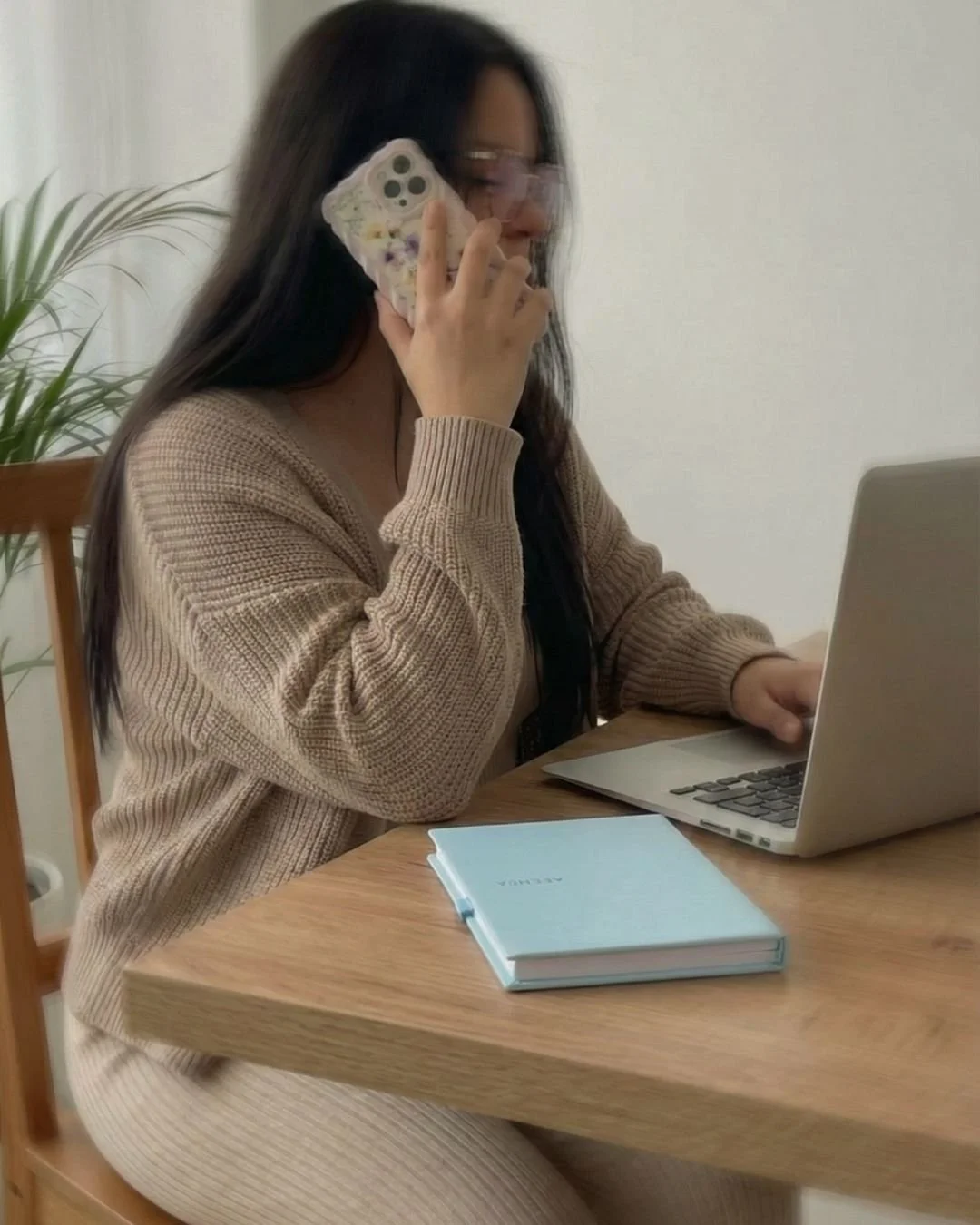 Una mujer con gafas claras, una chaqueta beige, usando una computadora portátil y hablando por teléfono, en una mesa de madera con una libreta azul.