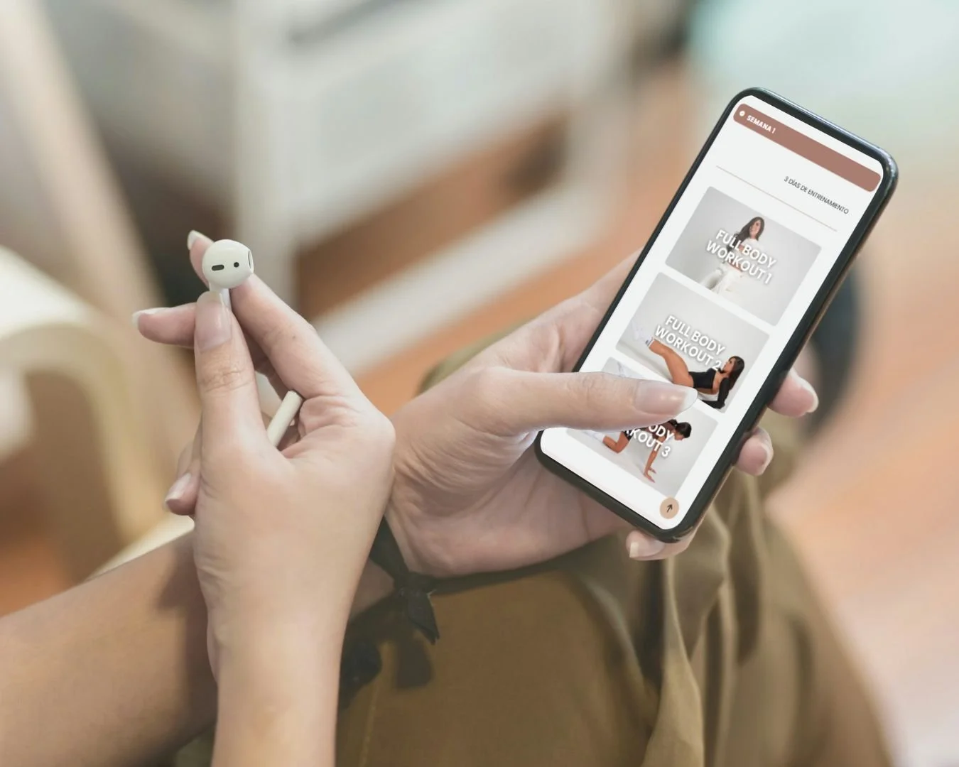 Persona sosteniendo un teléfono móvil y unos auriculares, viendo una aplicación de entrenamiento con videos de ejercicios en la pantalla.