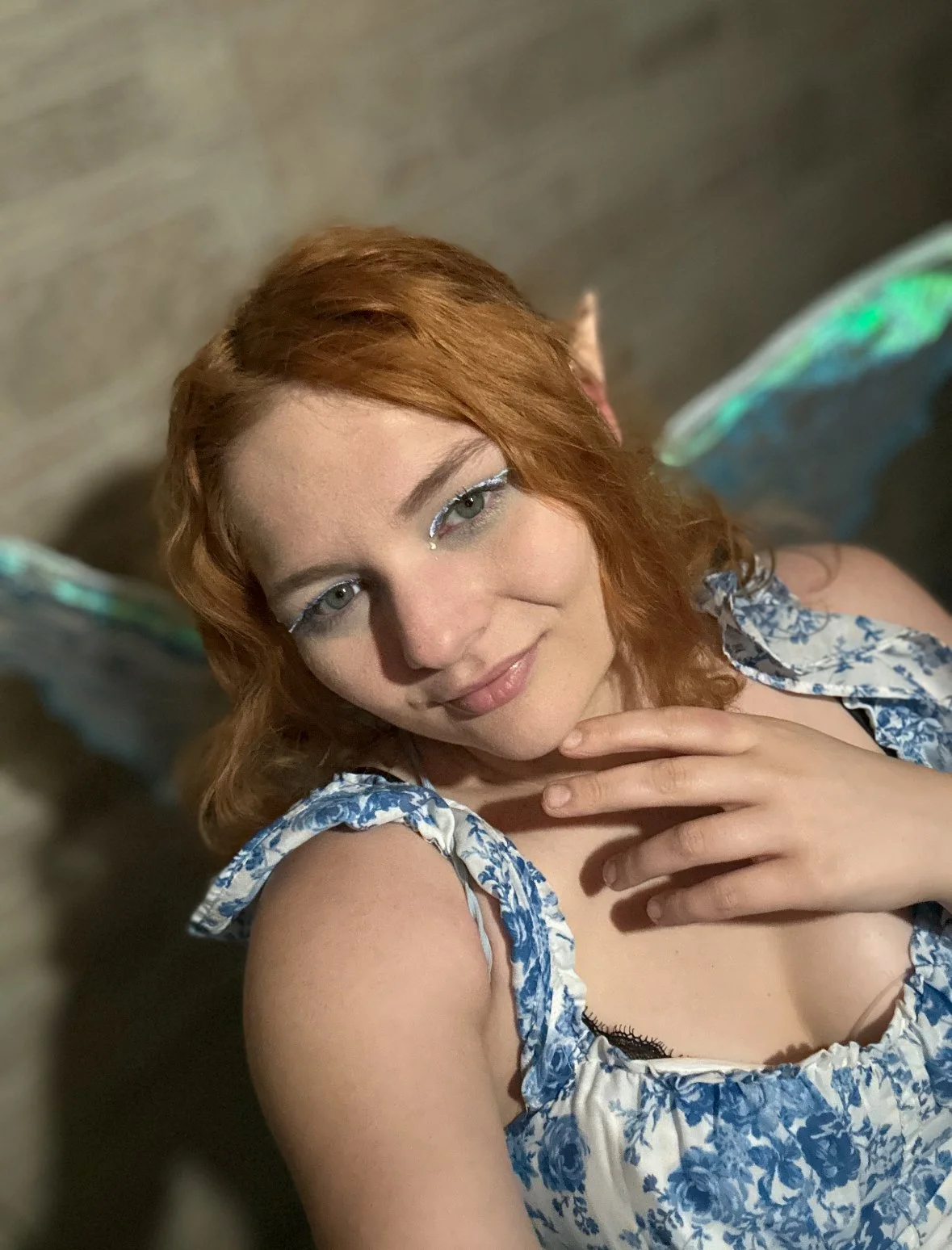 A woman with red hair and fairy wings, wearing a blue and white floral dress, posing indoors with a hand near her neck and smiling.