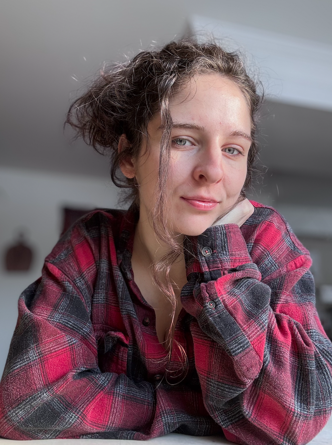 A young woman with curly brown hair, wearing a red and black plaid shirt, resting her head on her hand, looking at the camera with a slight smile, in a well-lit room.