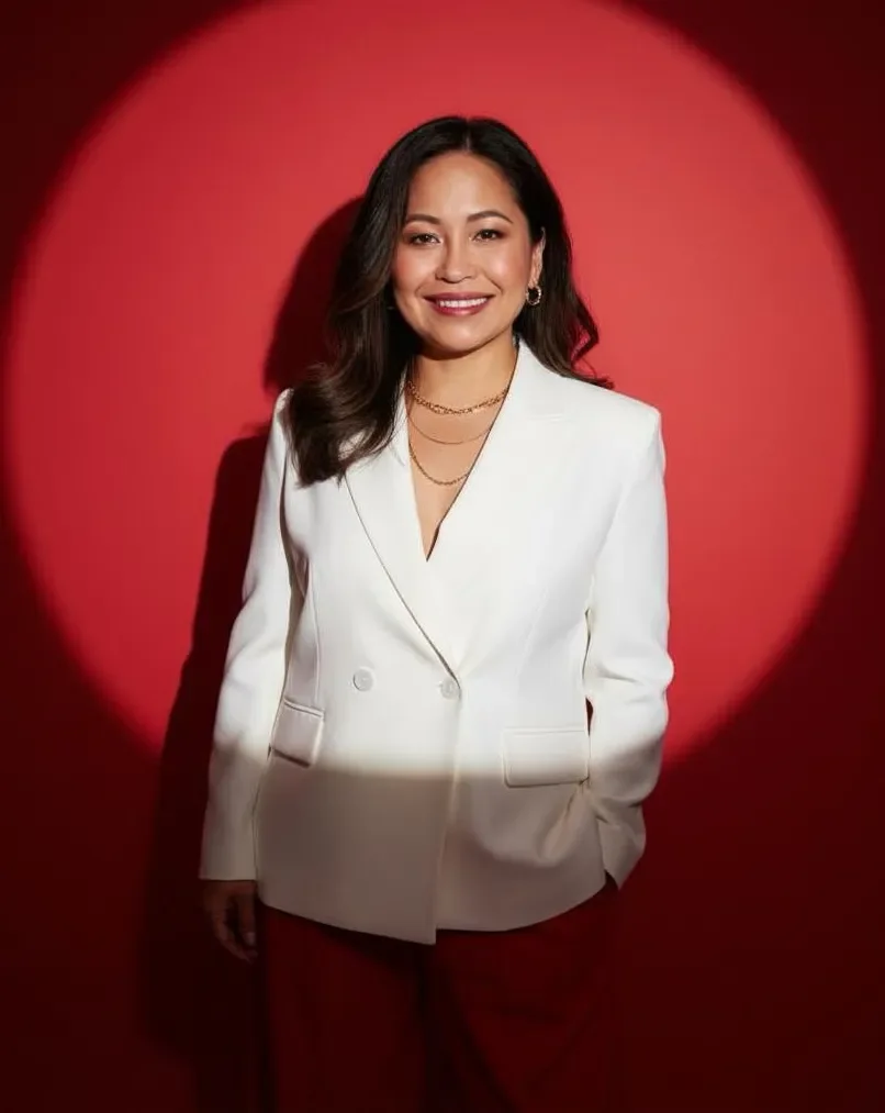 A woman in a white blazer and gold jewelry standing against a red background with a spotlight effect.