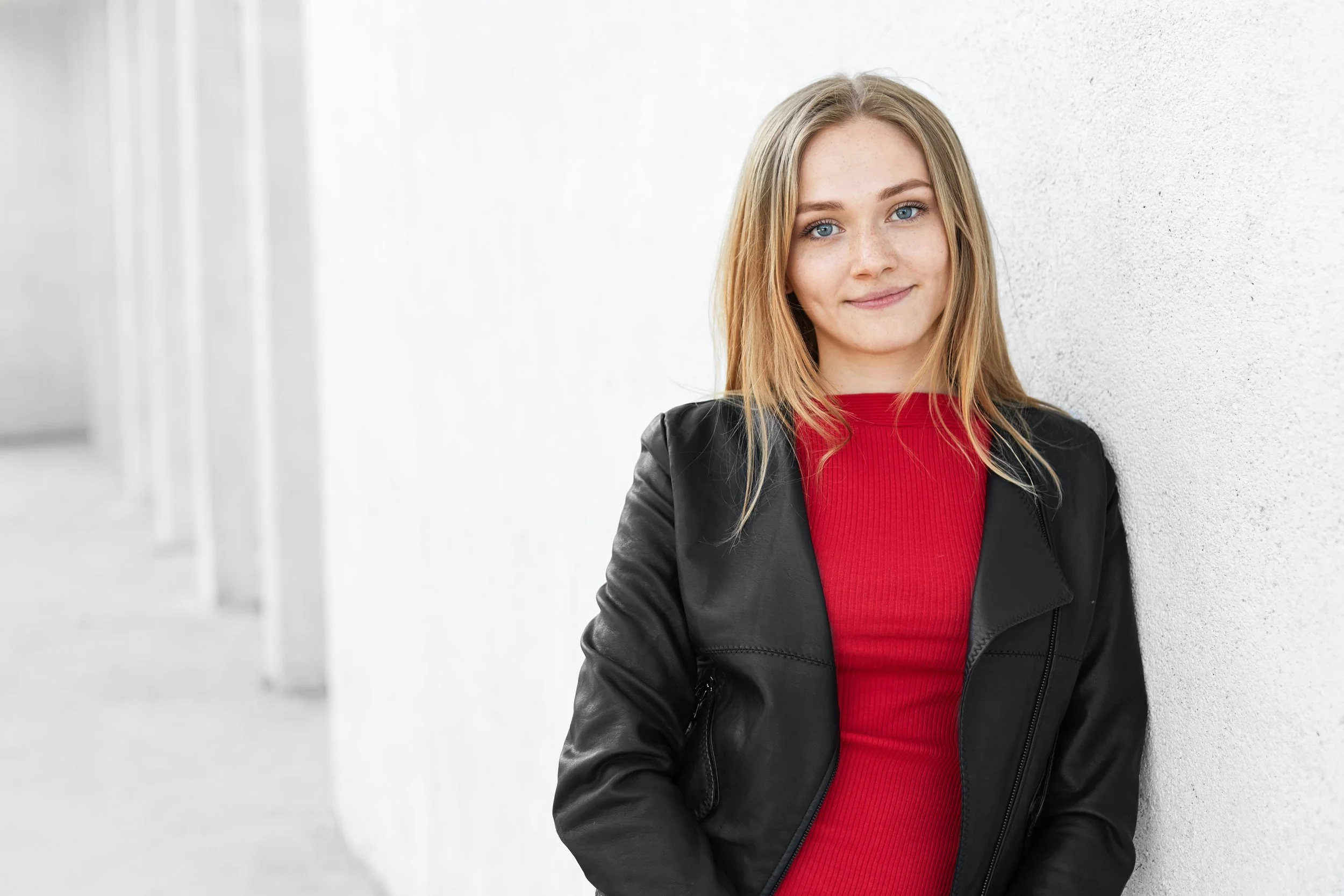 A young woman with blonde hair and blue eyes leaning against a white wall, wearing a red top and black leather jacket, smiling gently at the camera.