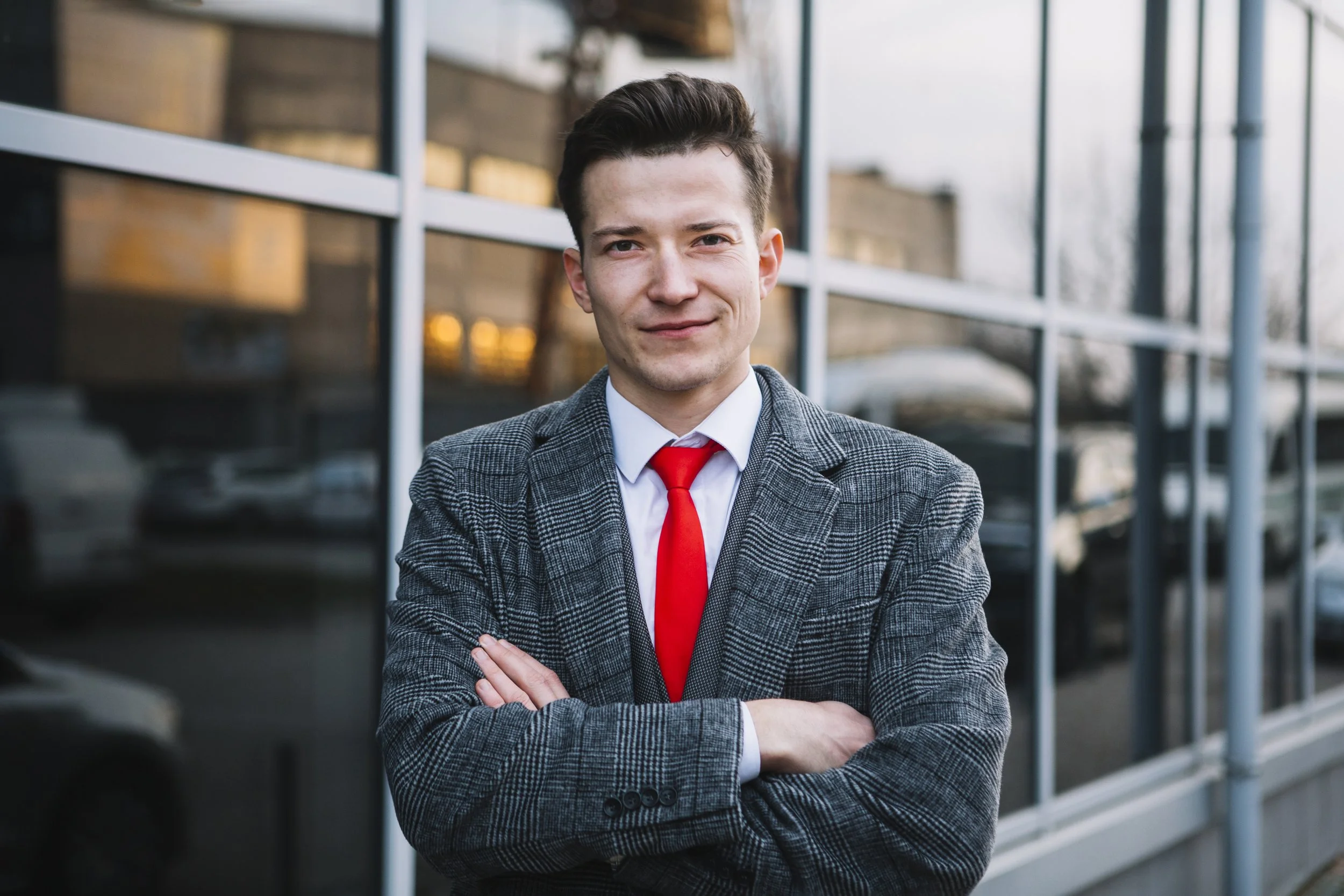 A young man dressed in a gray plaid suit with a white shirt and red tie standing outside office building with glass windows, arms crossed, smiling.