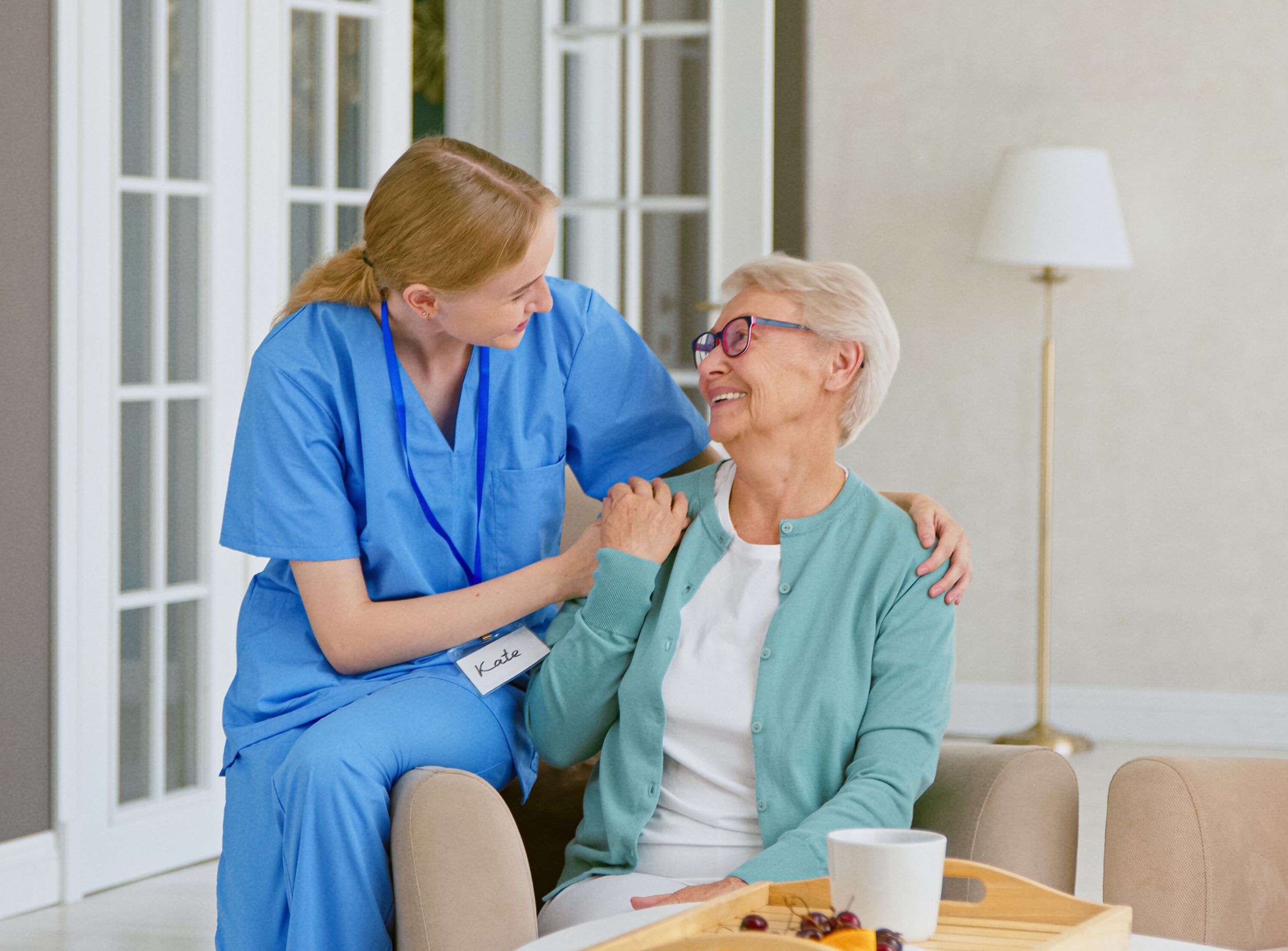 A healthcare worker in blue scrubs, wearing a name tag that reads 'Kate', is smiling and talking with an elderly woman in a green cardigan and glasses, sitting in a living room. The healthcare worker is gently holding the elderly woman's shoulder and both are smiling at each other.