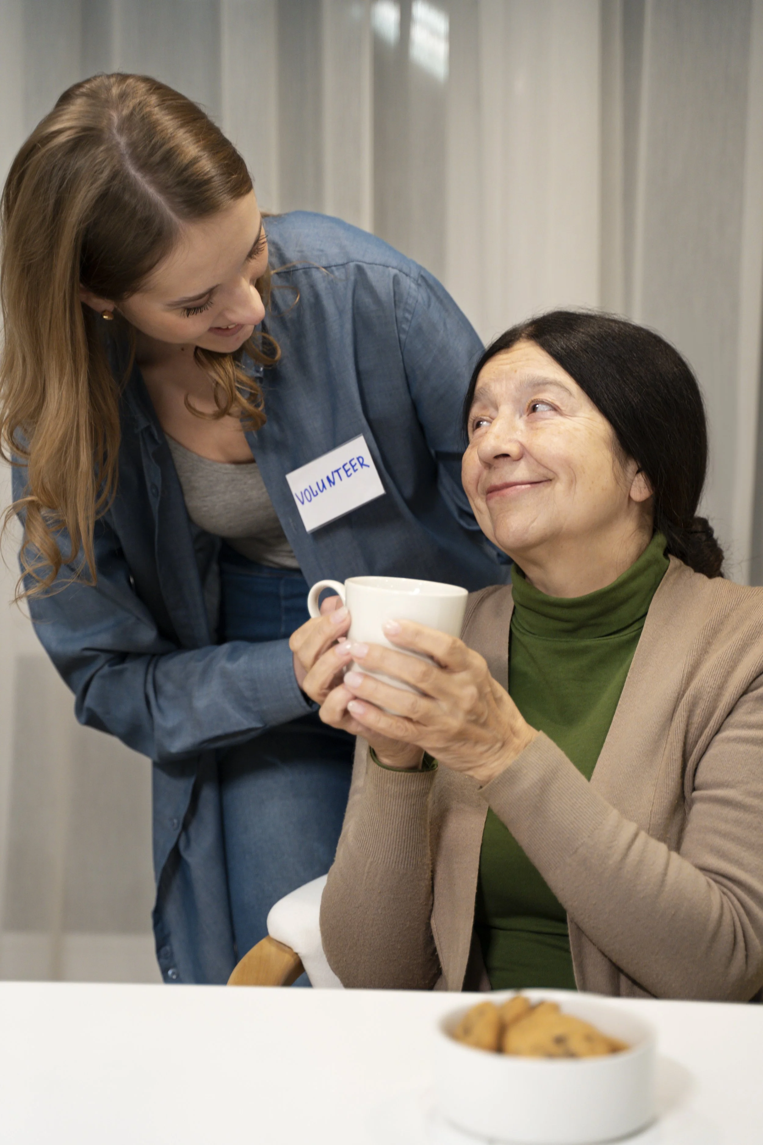 A young woman volunteer smiling and handing a mug to an elderly woman seated at a table, with a bowl of cookies in the foreground.