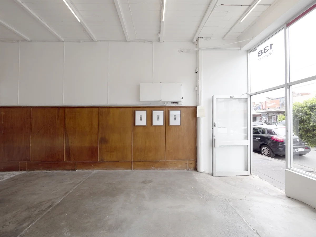 Interior of art gallery with a glass door and large front window, wooden paneling on the lower half of the wall, three framed pictures, artwork by artist Thomas Pendergast, and an exterior parking space visible through the glass.
